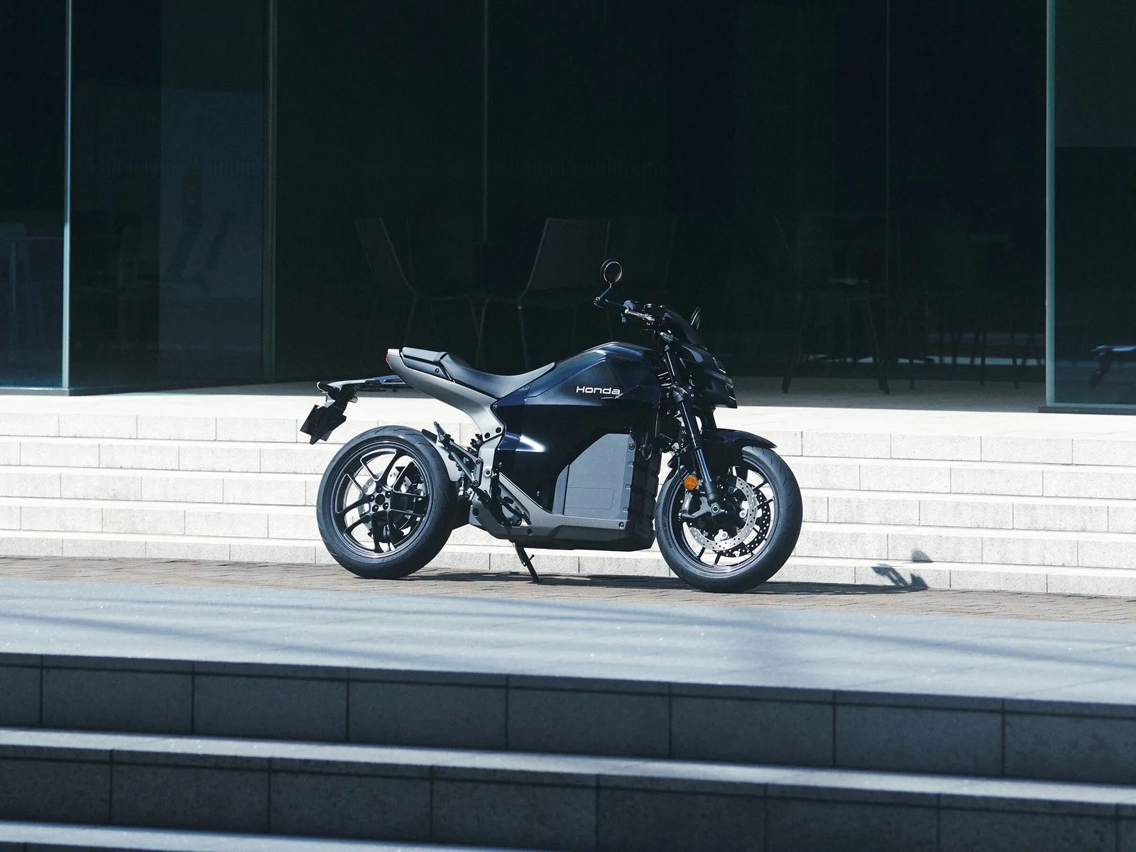 Black Honda electric motorcycle parked on a paved area in front of light-colored steps and glass windows.