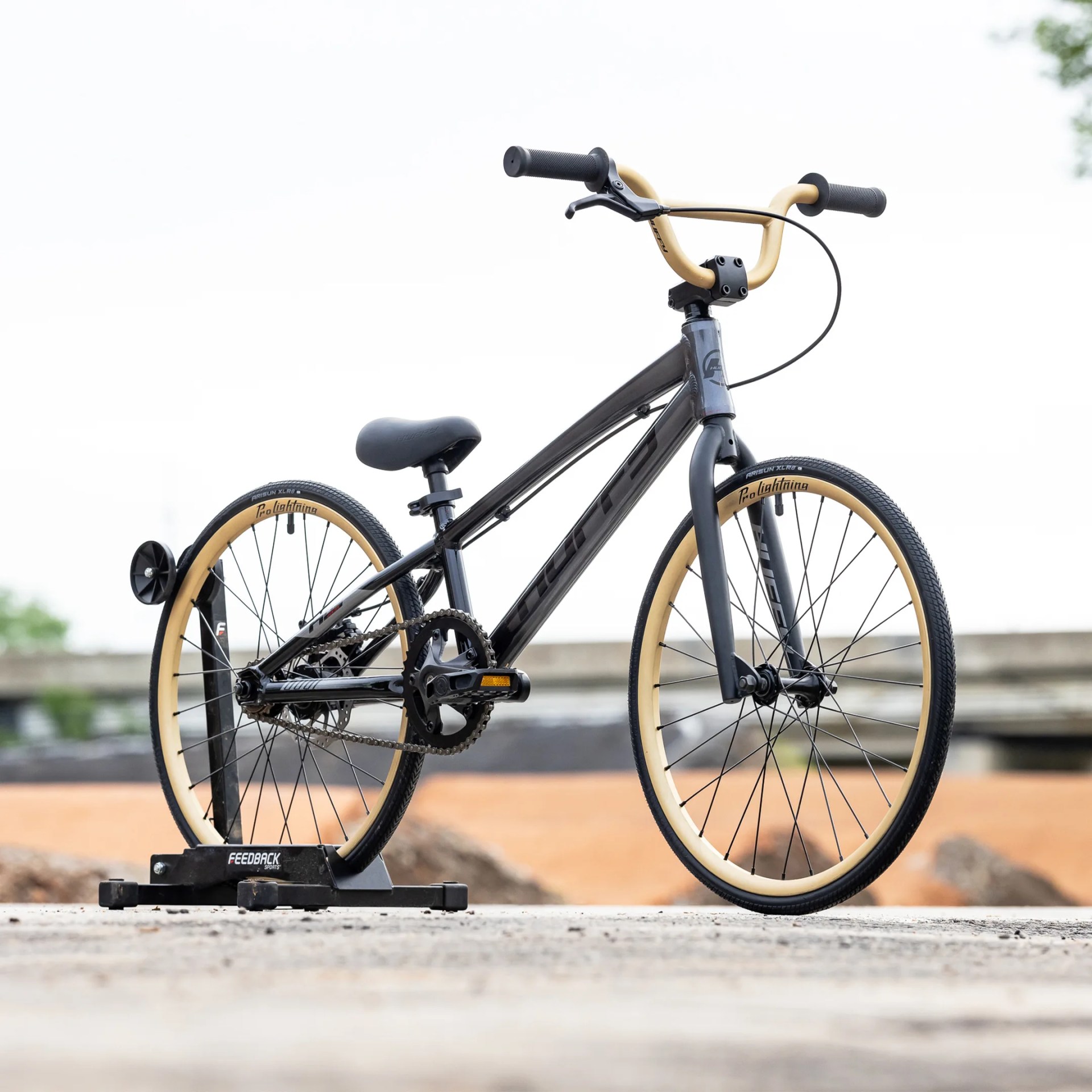 Black BMX bike with tan tires mounted on a black Feedback Sports bike stand outdoors.