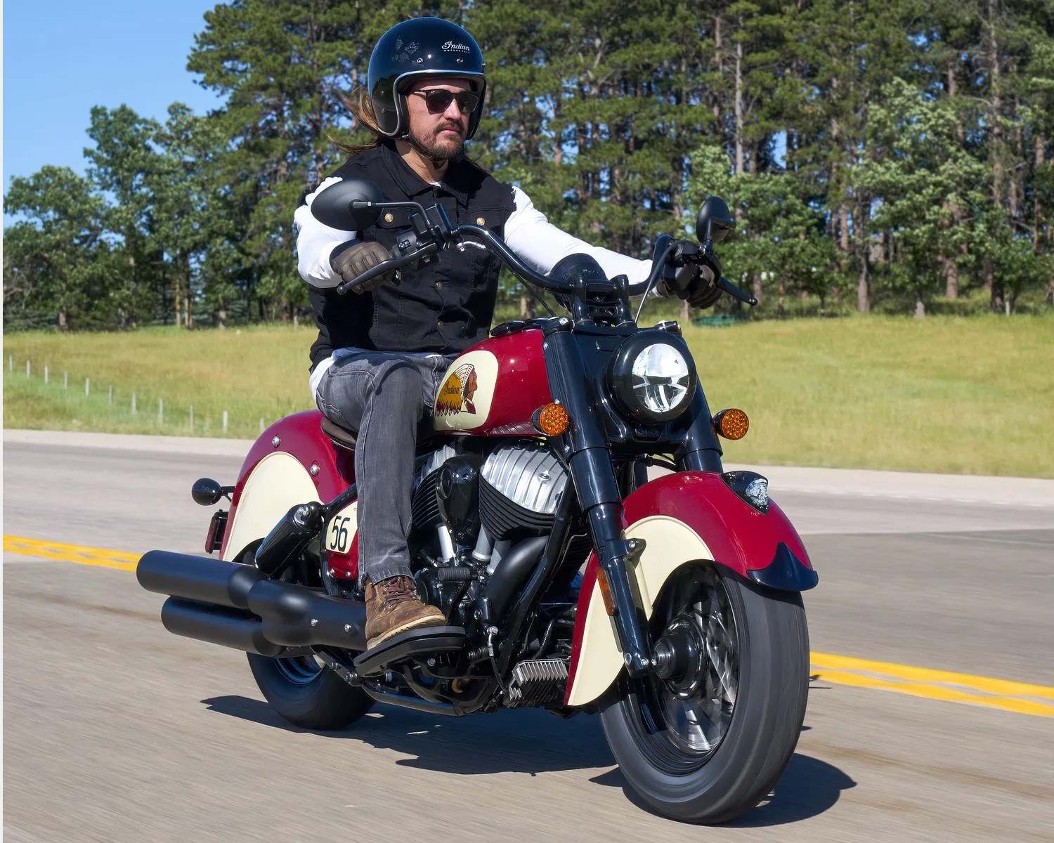 Man wearing a black helmet and vest riding a red and cream Indian motorcycle on a road with trees in the background.