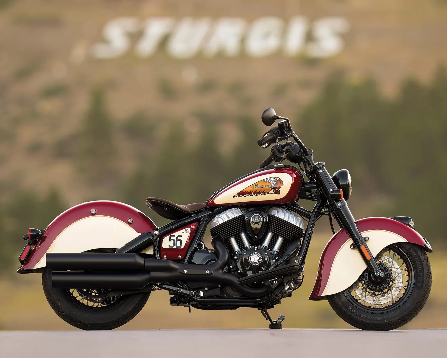 Red and cream Indian motorcycle with black engine and exhaust pipes parked outdoors with "STURGIS" sign blurred in background.