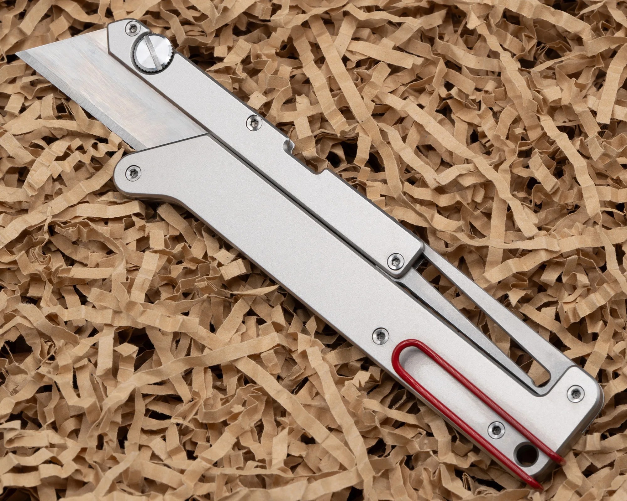 Silver metal folding utility knife with a red clip resting on shredded brown paper.
