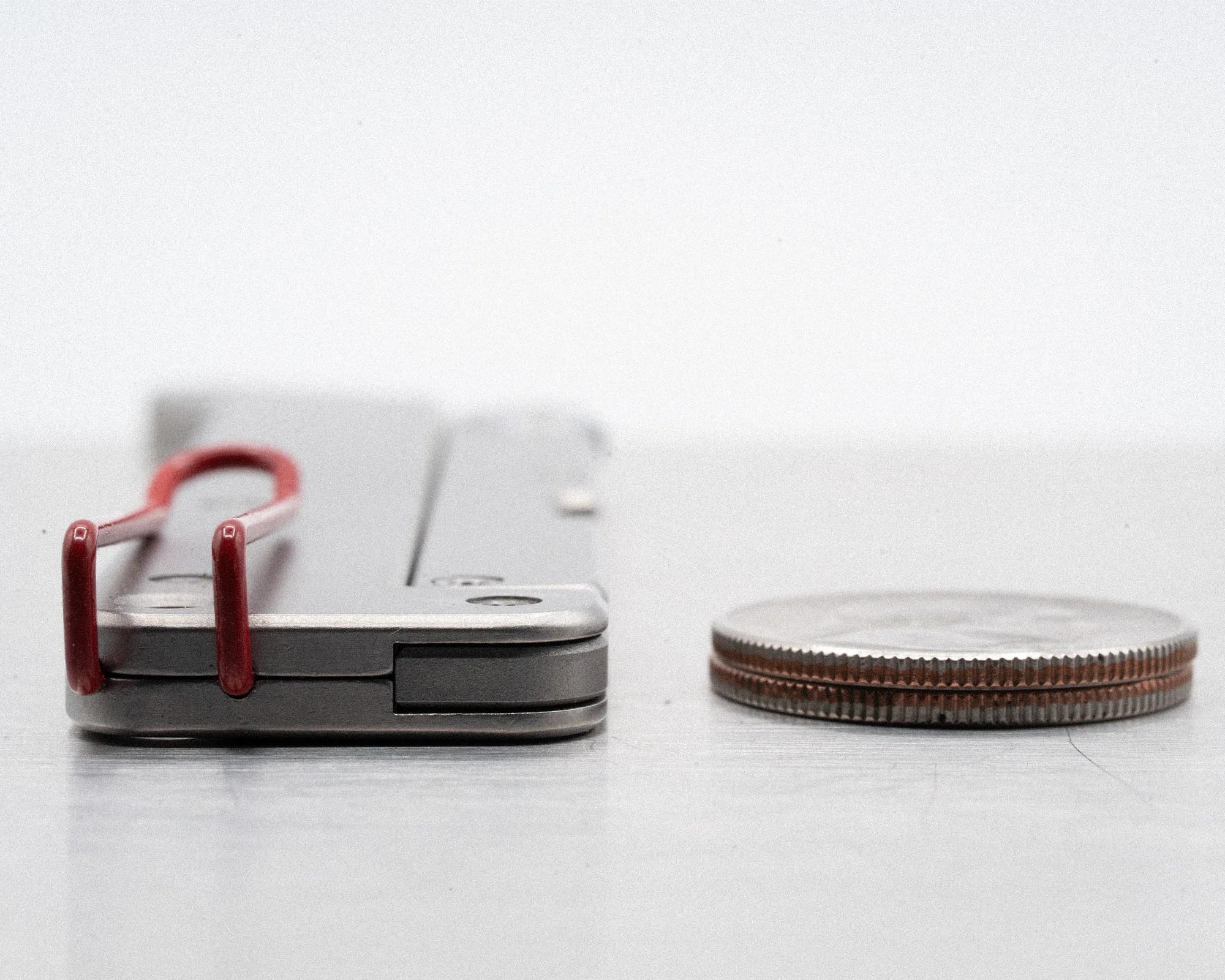 Stack of coins next to a closed gray metal money clip with red wire loops on a white surface.