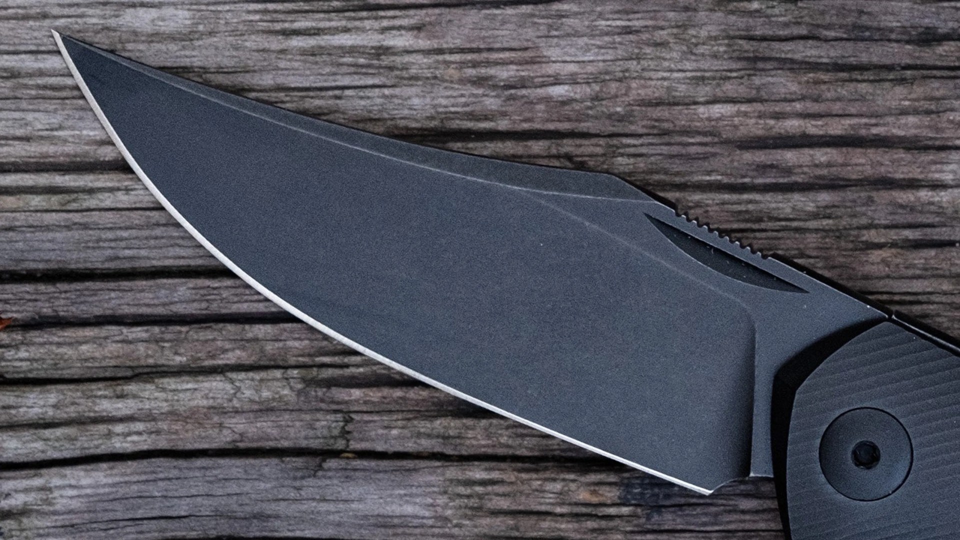 Close-up of a matte black folding knife blade on a weathered wooden surface.