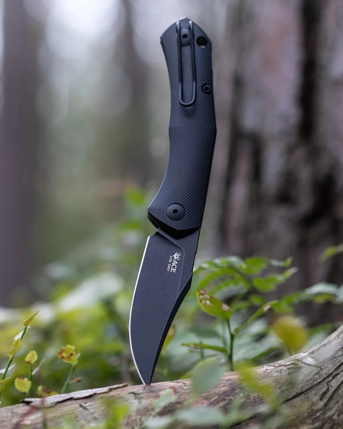 Black folding knife with textured handle and partially serrated blade stuck in a log outdoors.