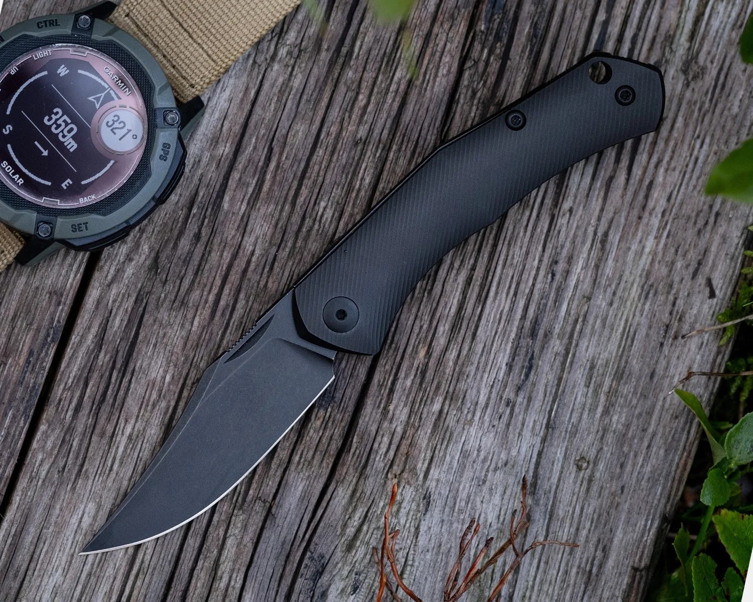 Black folding knife with textured handle on weathered wooden surface next to a wristwatch.
