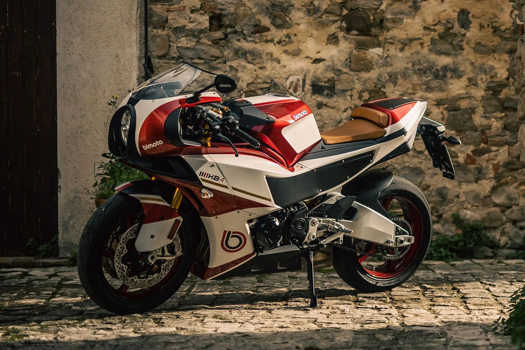 Red and white Bimota KB4 sport motorcycle with tan seat parked on cobblestone near stone wall.