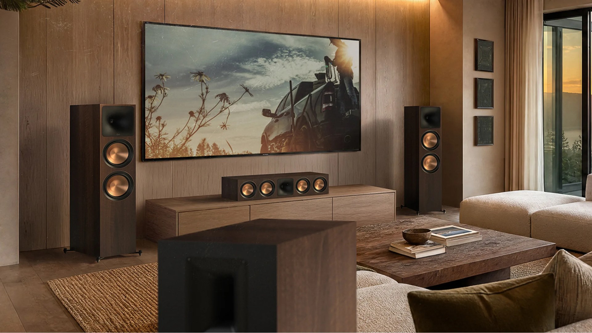 Living room with a wall-mounted flat-screen TV, two tall floor-standing speakers with copper cones, a center speaker on a wooden cabinet, and a beige sectional sofa.