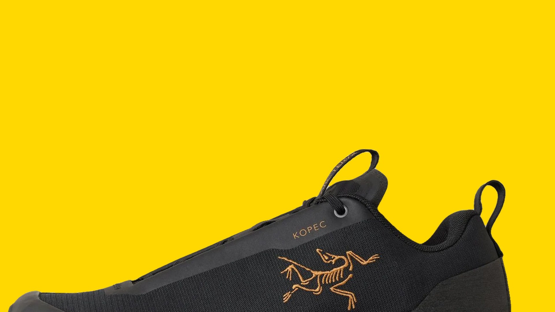 Black KOPEC sneaker with orange skeletal bird logo and pull tabs on yellow background.