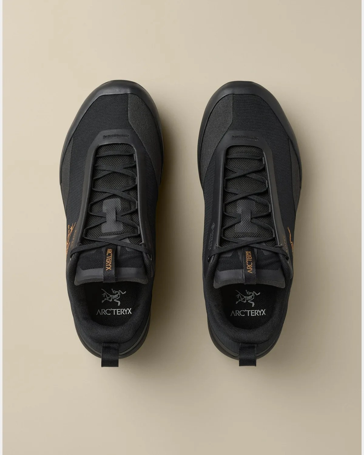 Pair of black Arc'teryx sneakers with Gore-Tex and orange logo details on a beige background.