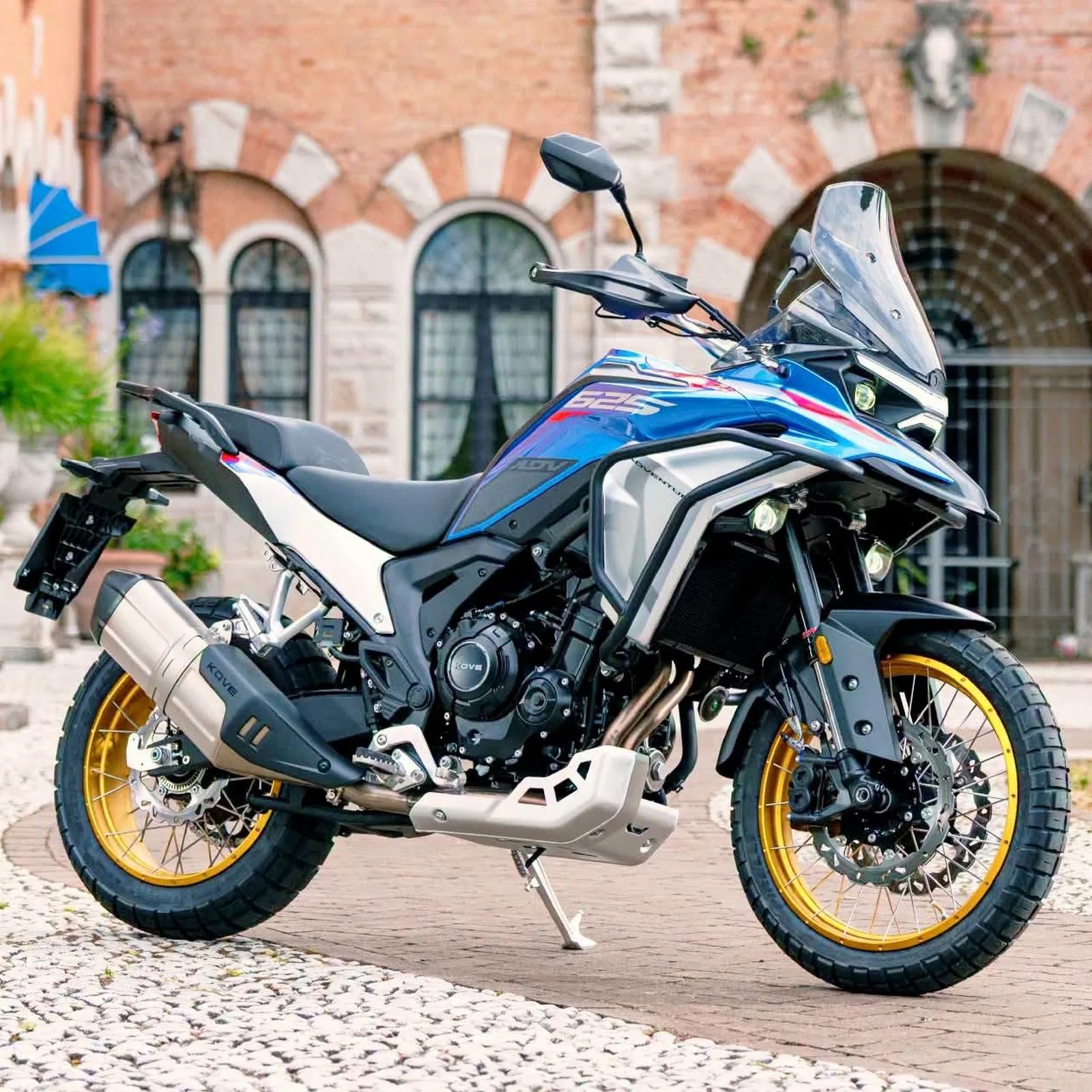 Blue and white adventure motorcycle with gold rims parked on a cobblestone street in front of a brick building.