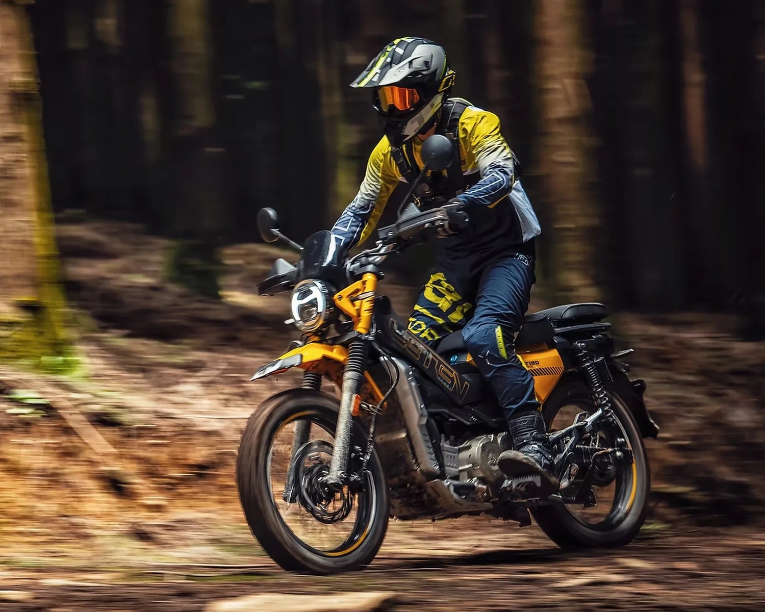 Person wearing yellow and blue riding gear and helmet on a yellow and black off-road motorcycle in a forest.
