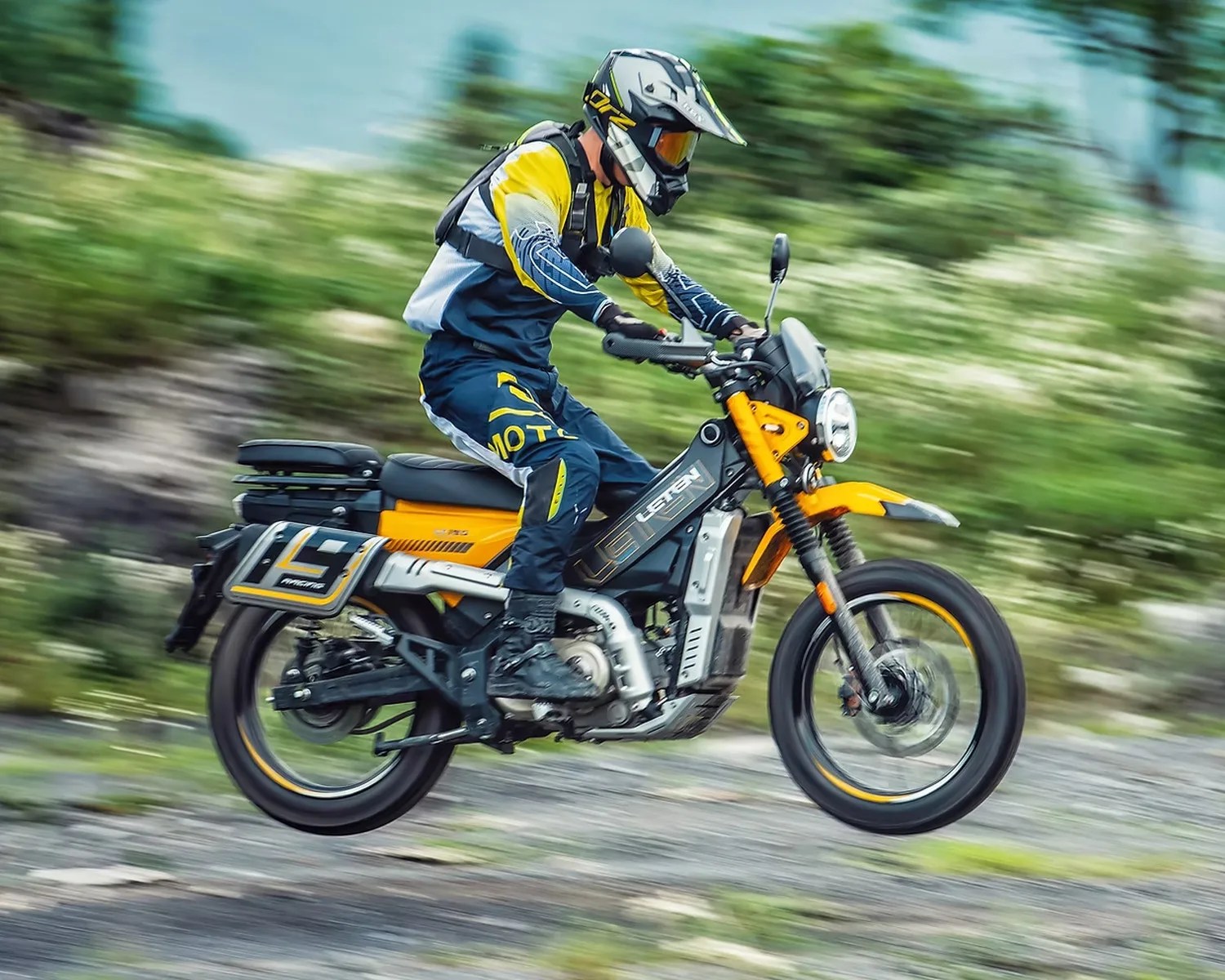Person wearing a yellow and blue riding suit and helmet riding a yellow and black electric motorcycle on a gravel path.