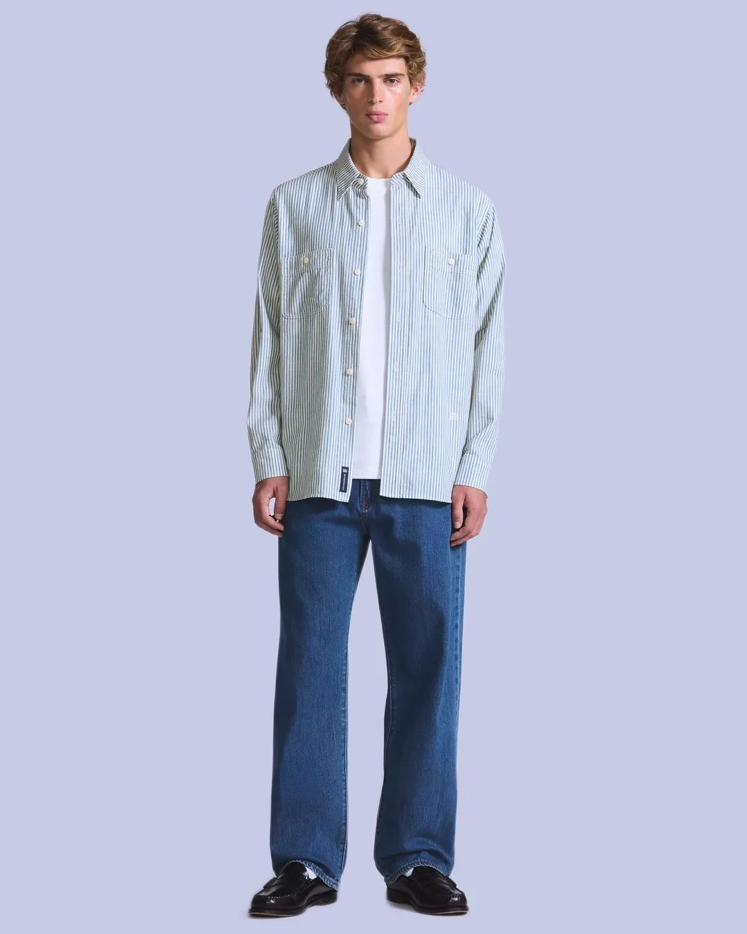 Man wearing light blue and white vertical striped button-up shirt over a white T-shirt, blue jeans, and black shoes.