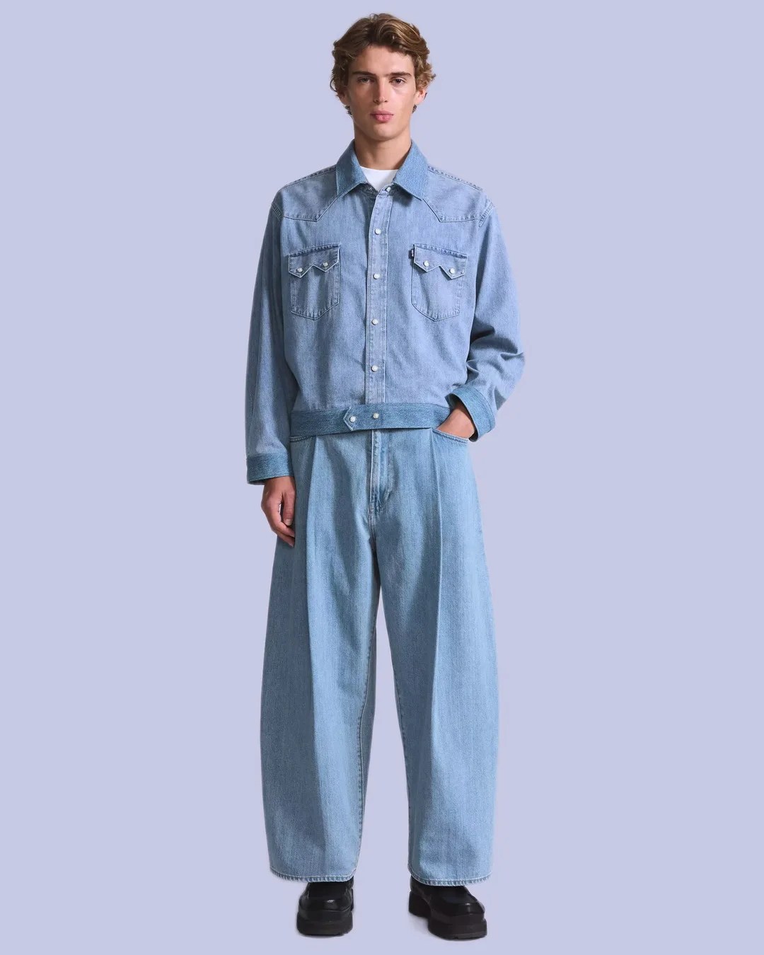 Man wearing light blue denim jacket with snap buttons and matching wide-leg jeans against a plain background.