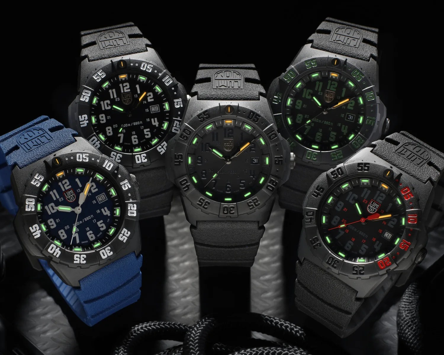 Five Luminox wristwatches with black cases and luminous green and orange markers, one with a blue strap, displayed on a dark textured surface.