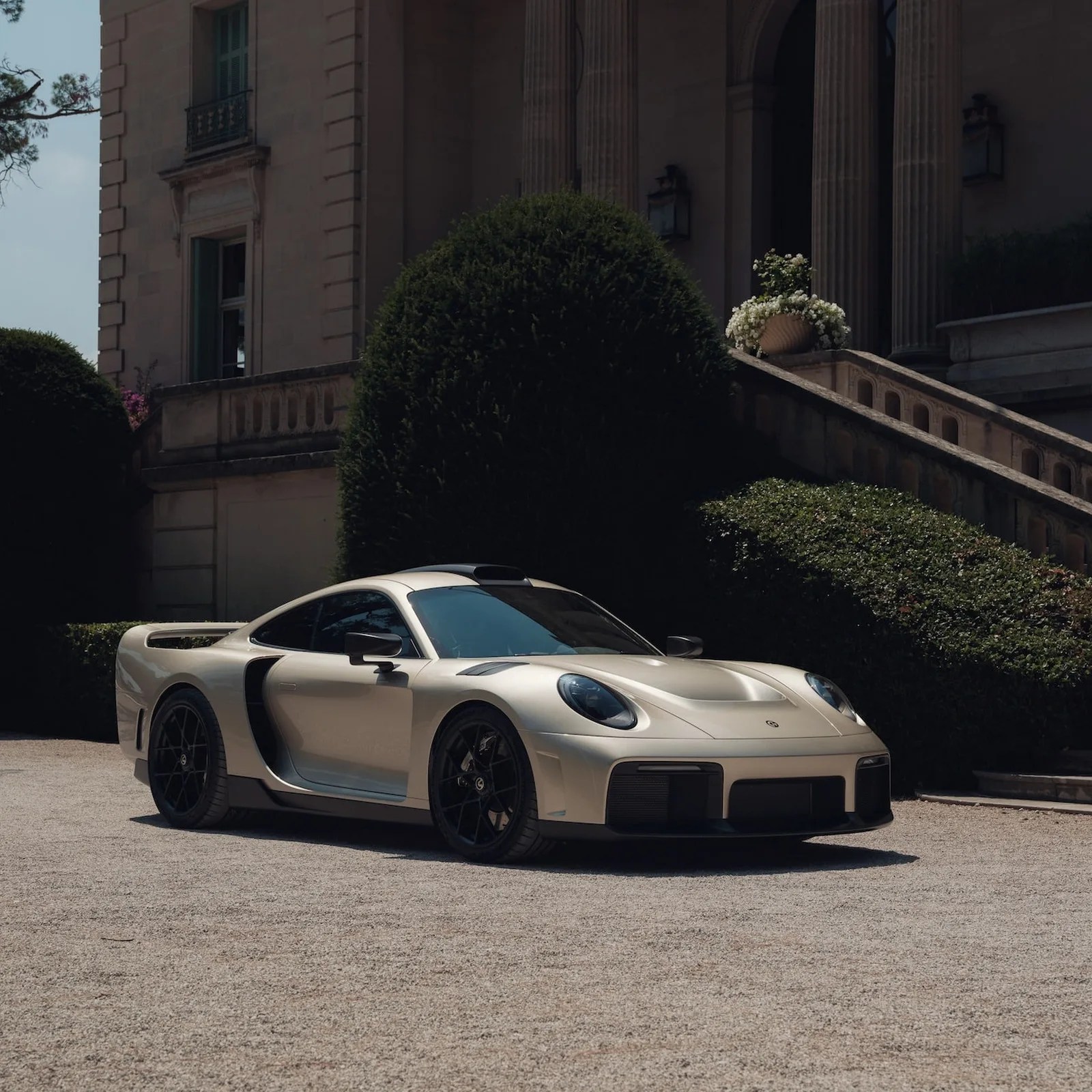 Champagne-colored sports car with black wheels parked on gravel in front of a classical building with large bushes.