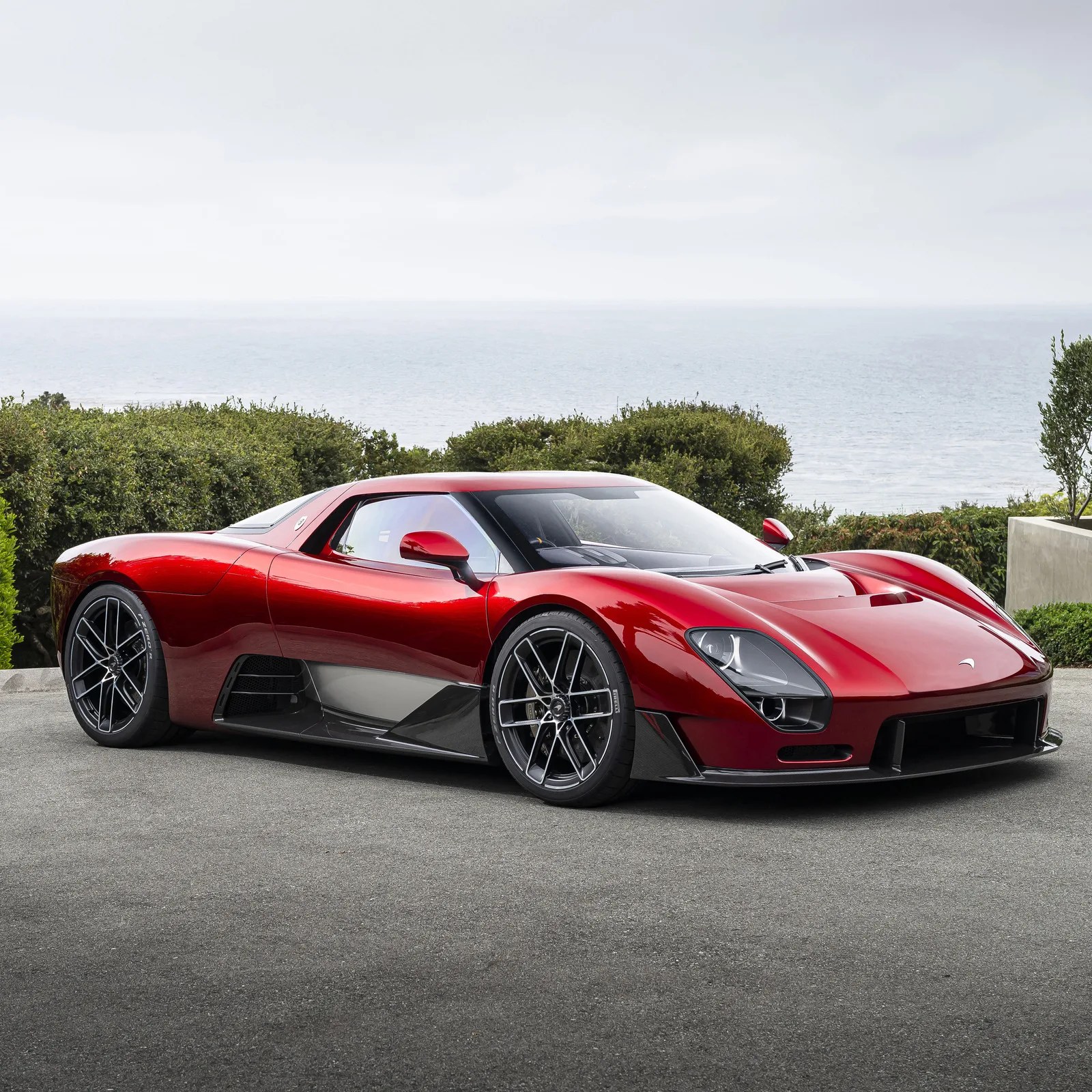 Shiny red McLaren Speedtail hypercar parked on a driveway with ocean and greenery in the background.