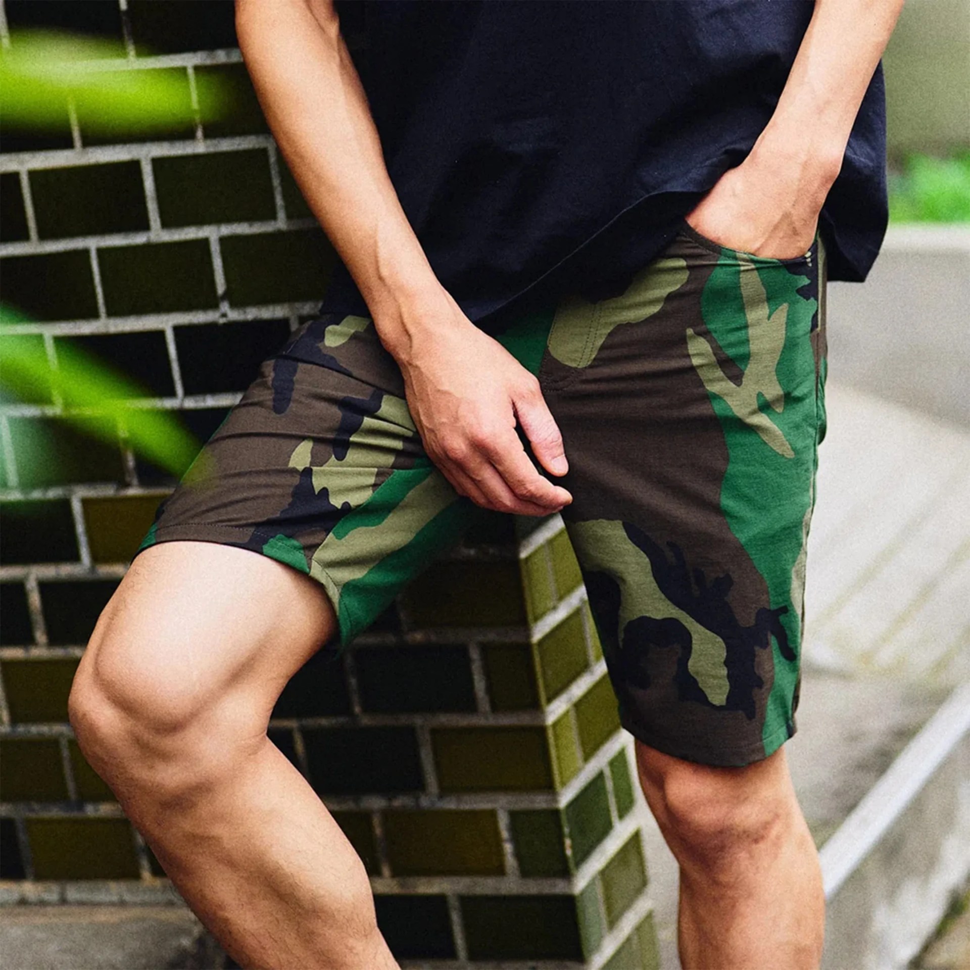 Person wearing green and brown camouflage shorts and a black shirt, with one hand in the pocket.