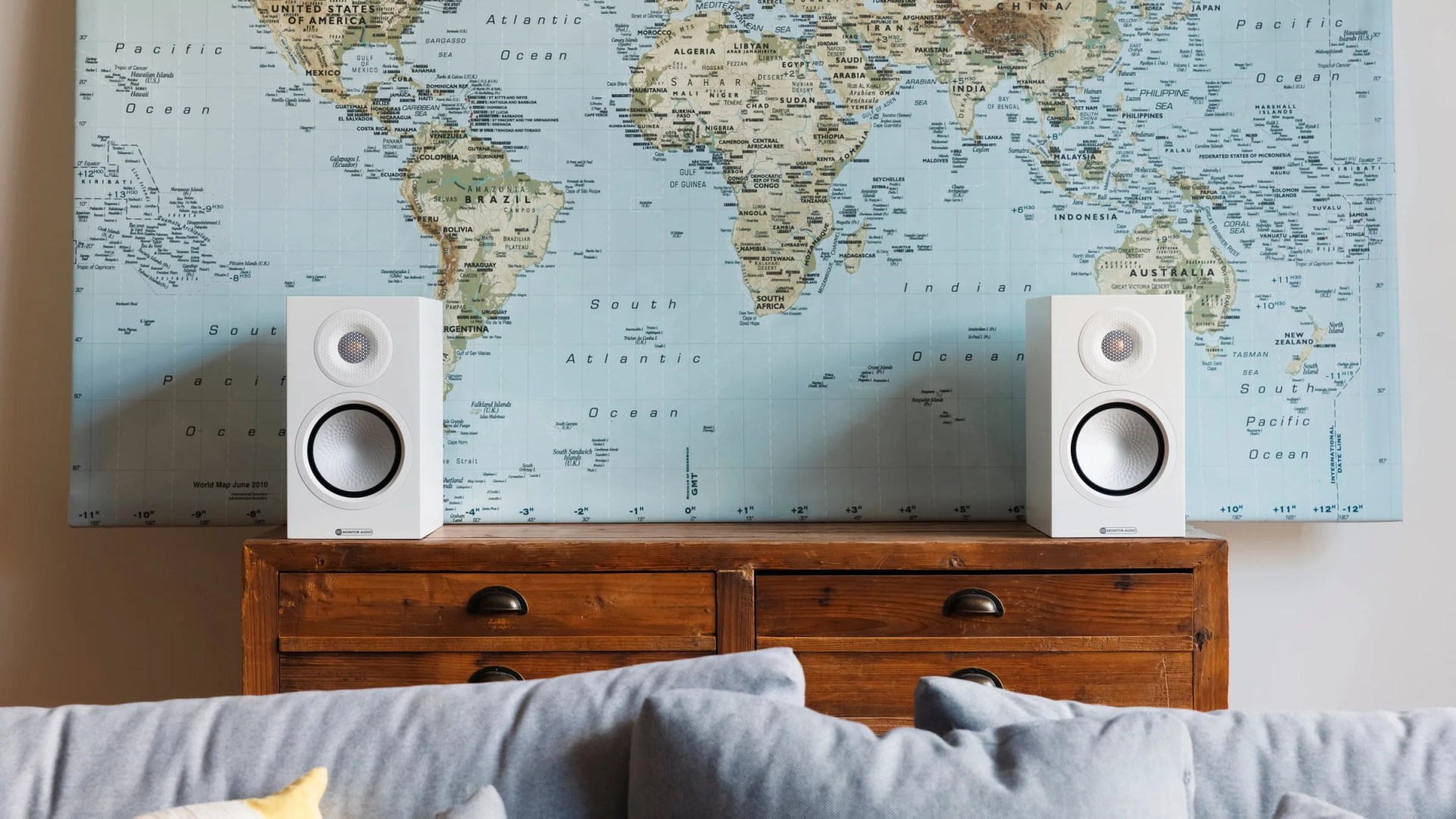 Pair of white bookshelf speakers on a wooden dresser with a world map on the wall behind.