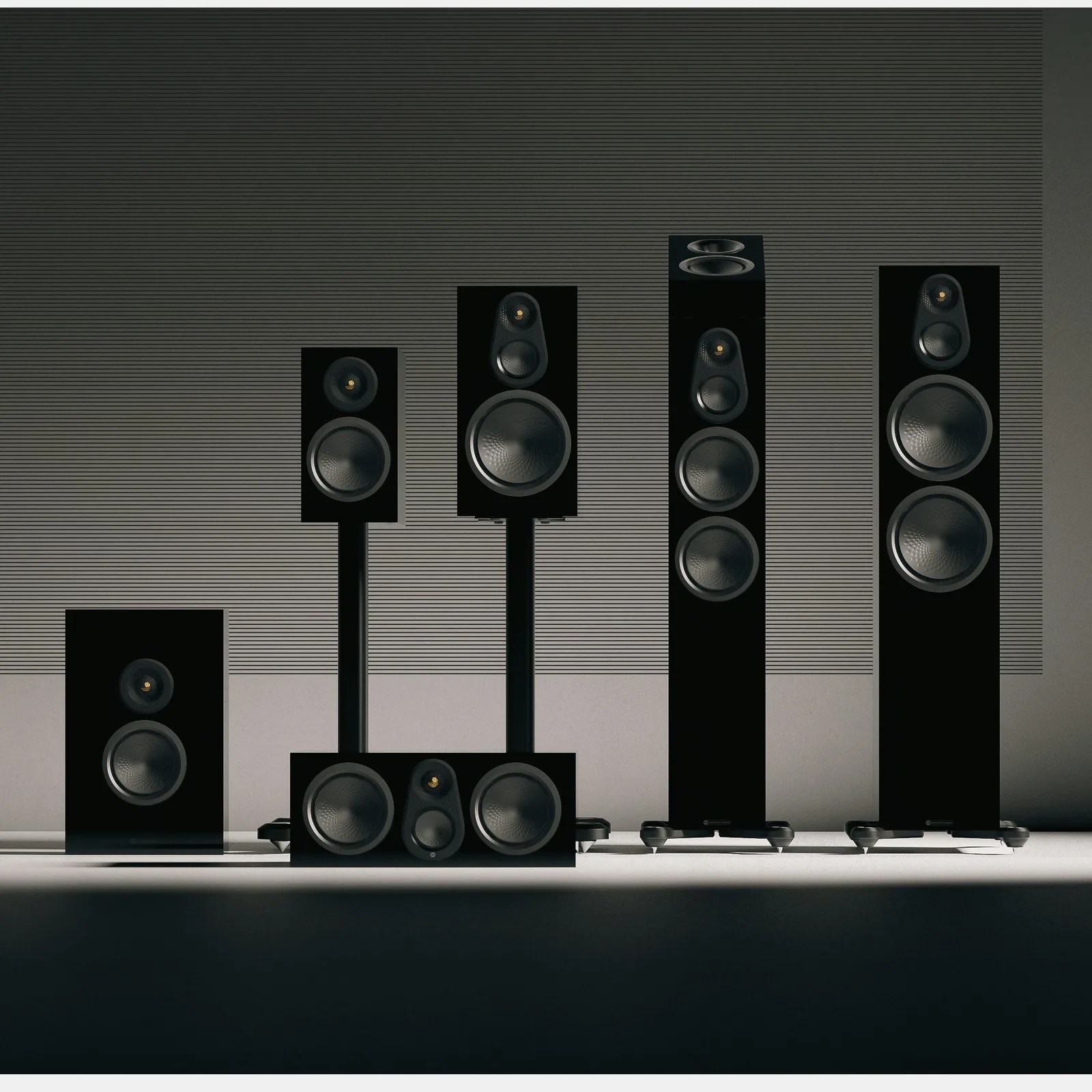 Five black modern speakers of varying sizes arranged in a row against a striped gray wall.