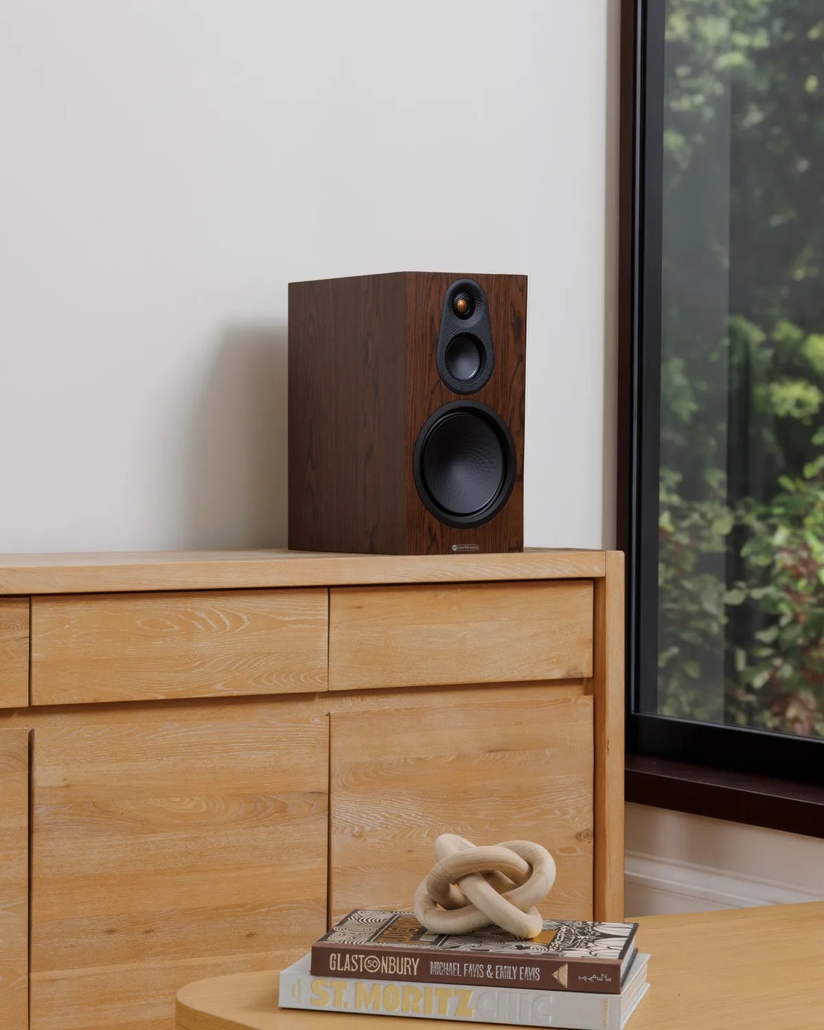 Wooden bookshelf speaker with black drivers on a light wood cabinet next to a window.