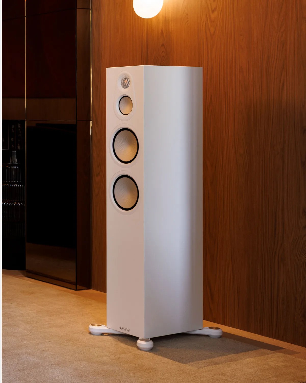 Tall white floor-standing speaker with three circular drivers on a beige carpet in front of a wooden wall.