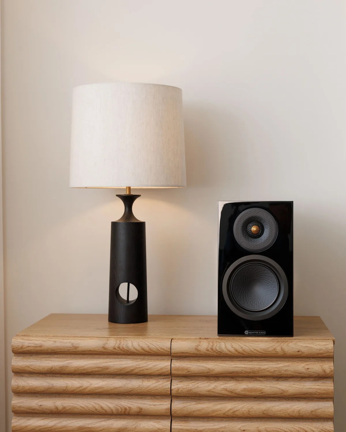 Black table lamp with a white cylindrical shade and a black speaker with gold accents on a wooden dresser.