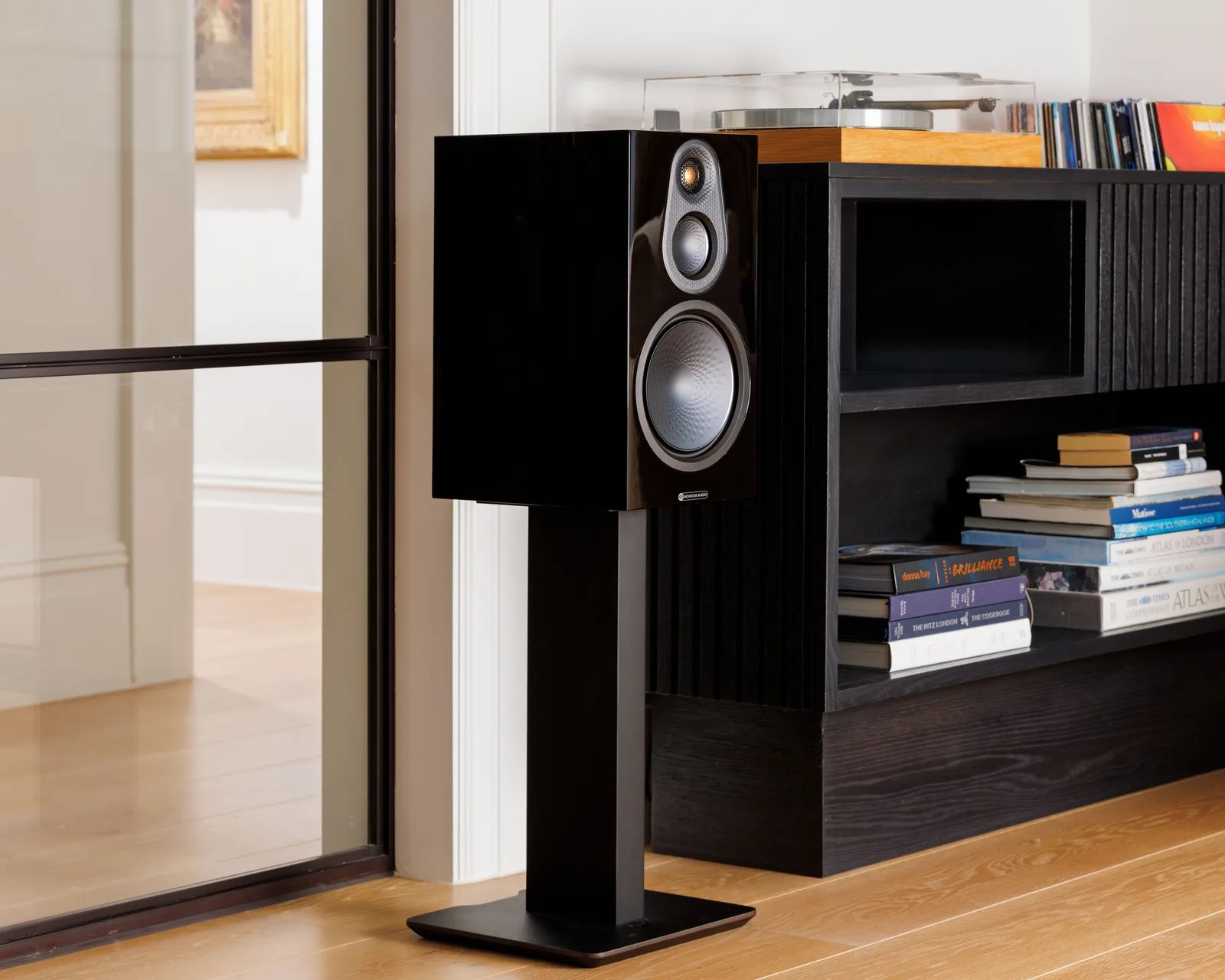 Glossy black Monitor Audio speaker on a black stand next to a black shelf with stacked books and a turntable.