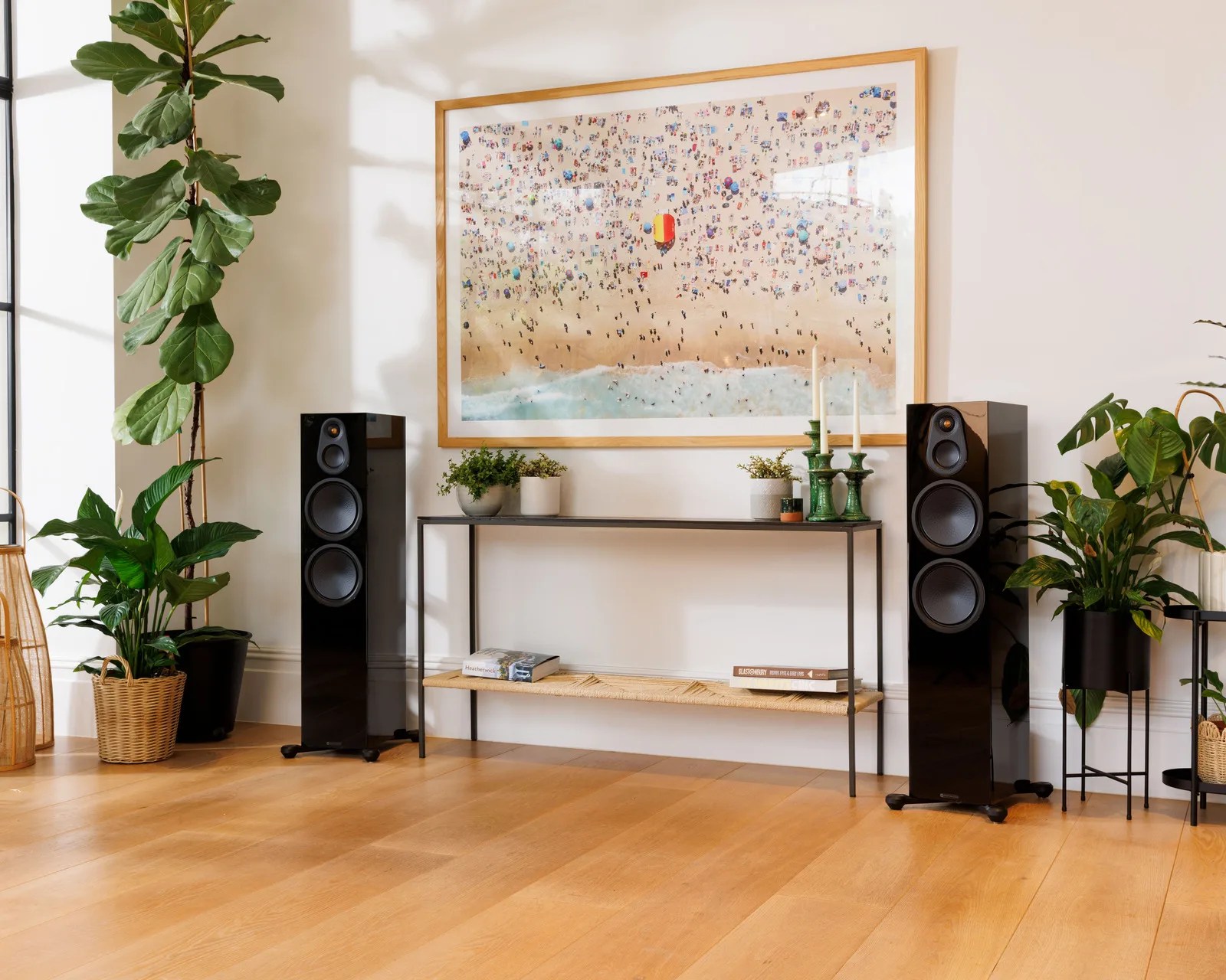 Black floor-standing speakers flanking a slim black metal console table with plants, candles, and books under a large framed aerial beach photo.