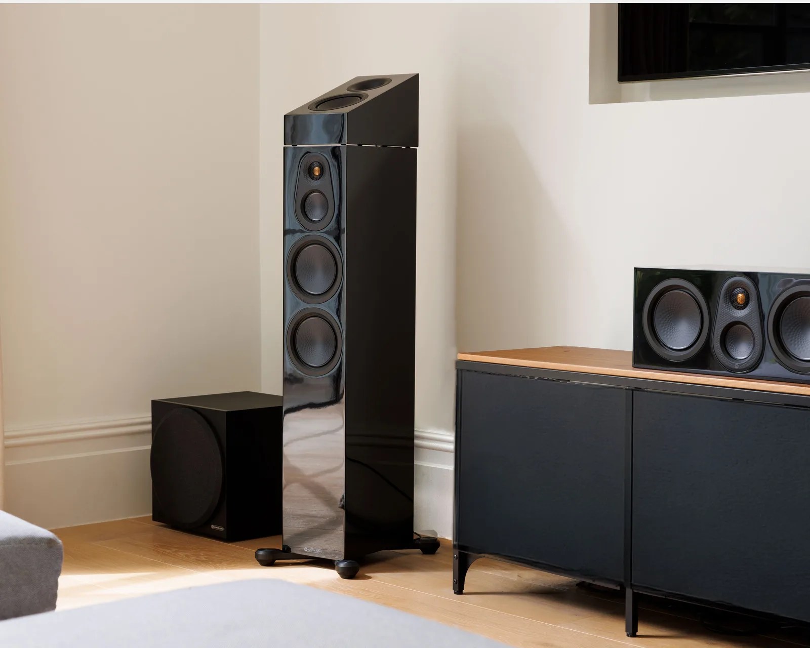 Glossy black floor-standing speaker with multiple drivers next to a black subwoofer and a black cabinet with a center speaker on top.