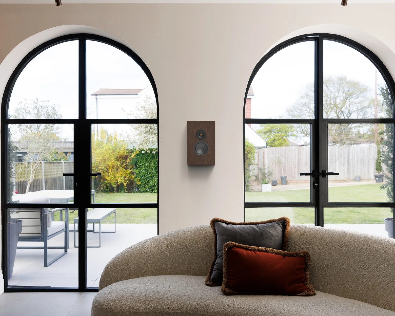 Curved beige sofa with two pillows in front of a wall-mounted wooden speaker between two large arched black-framed windows.