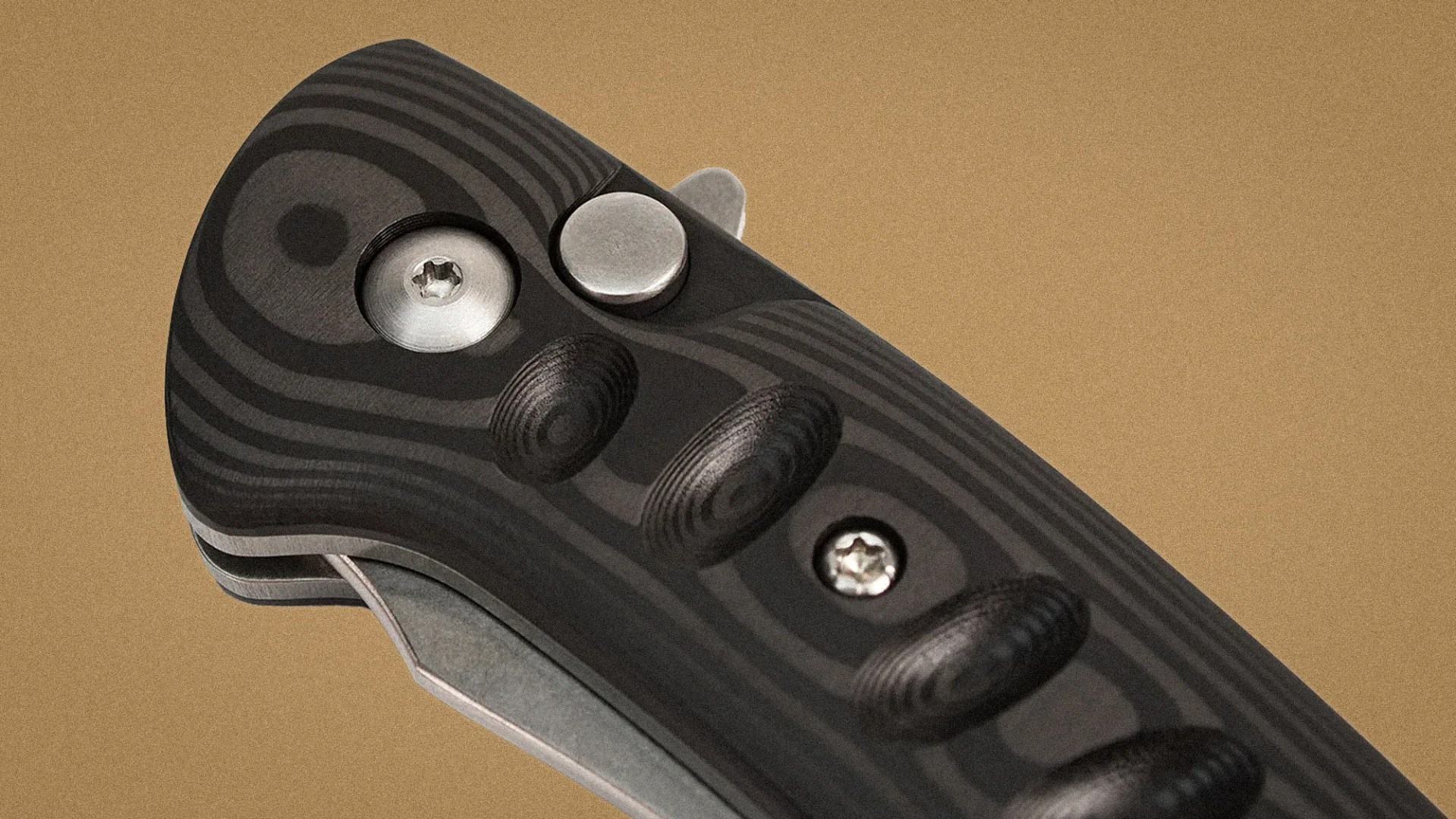 Close-up of a black and gray textured folding knife handle with metal screws and a button on a tan background.