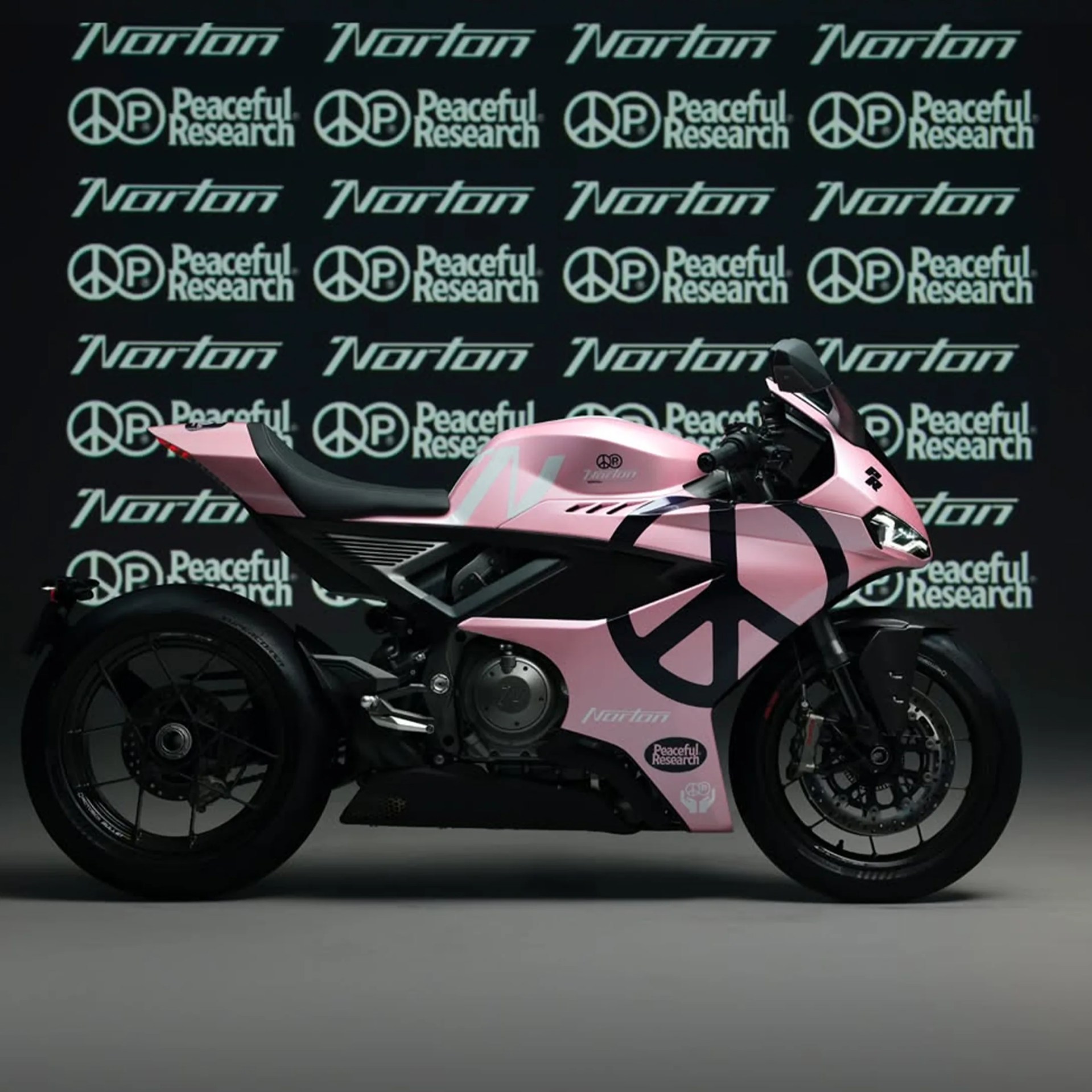 Pink Norton motorcycle with black peace symbol graphics and logos, set against a backdrop of repeating Norton and Peaceful Research logos.