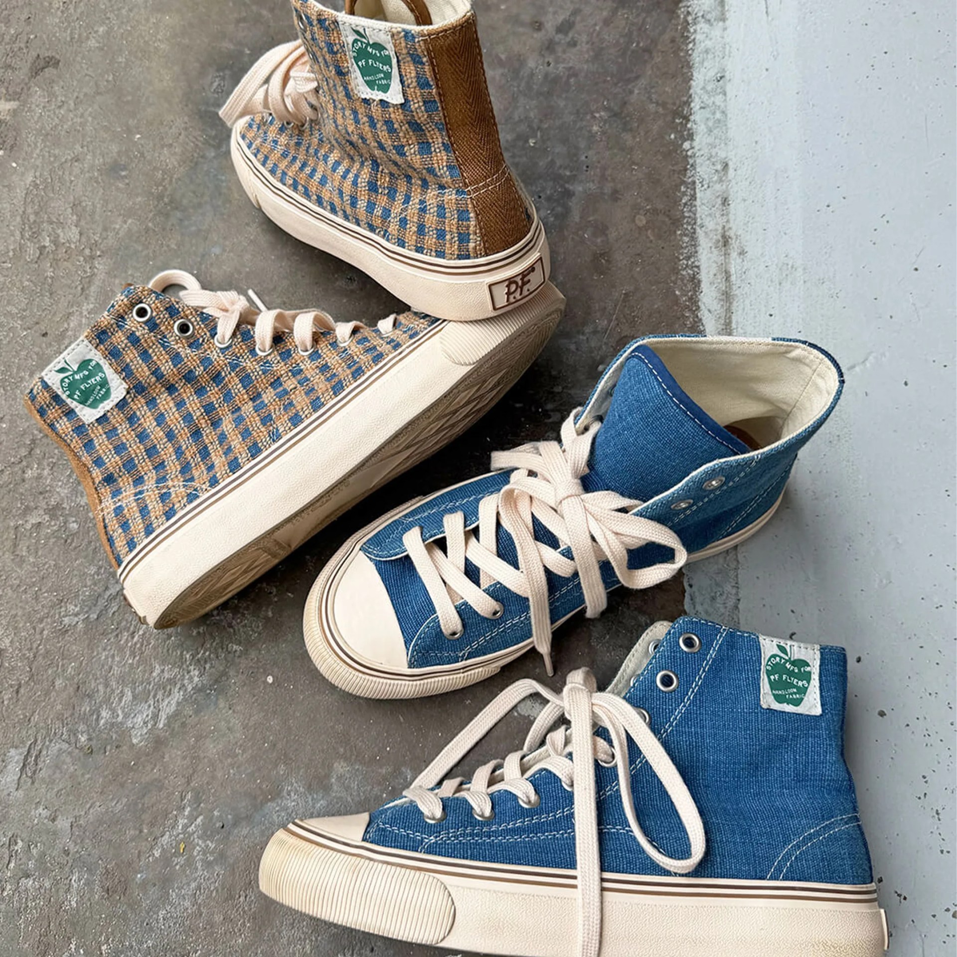 Two pairs of high-top canvas sneakers, one in blue denim with cream laces and soles, the other in brown and blue checkered fabric with cream soles, on a concrete floor.