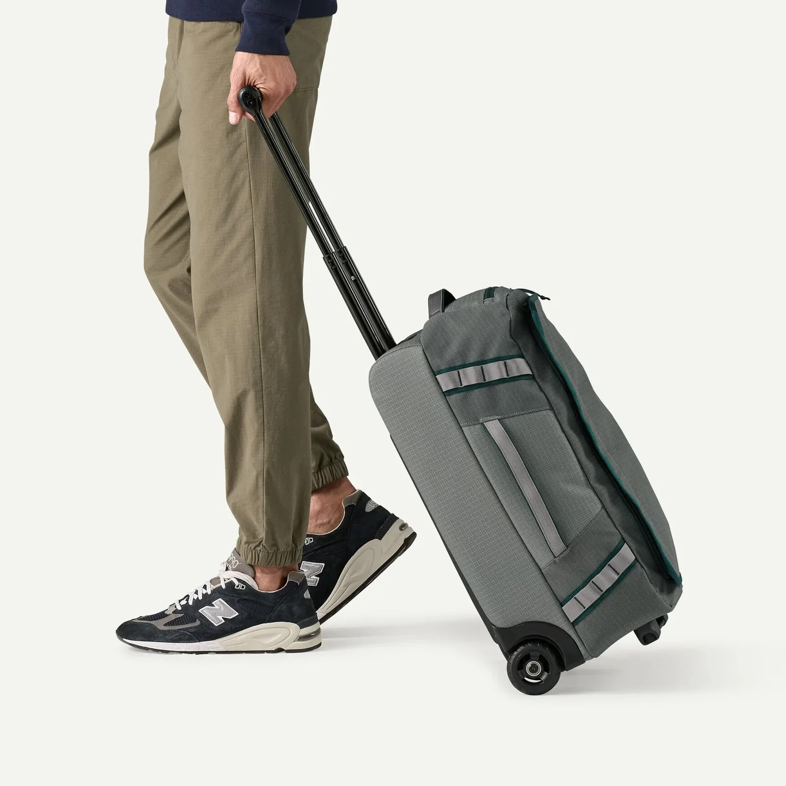 Person wearing olive jogger pants and black New Balance sneakers pulling a gray rolling suitcase with black handle.