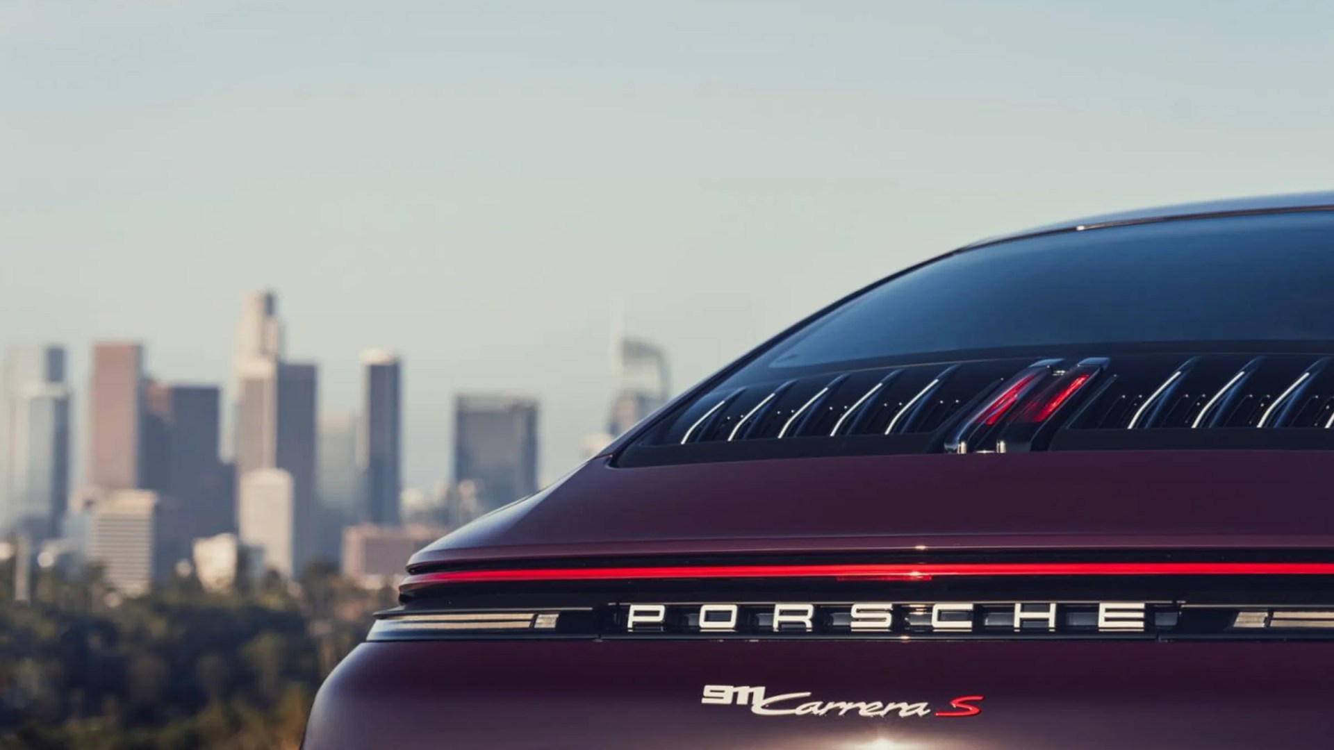 Rear view of a dark red Porsche 911 Carrera S with city skyline in the background.