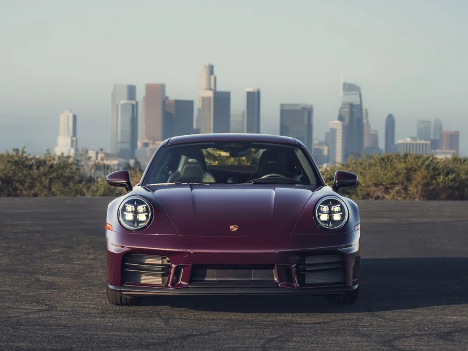 Front view of a dark purple Porsche sports car with LED headlights on an asphalt surface and a city skyline in the background.