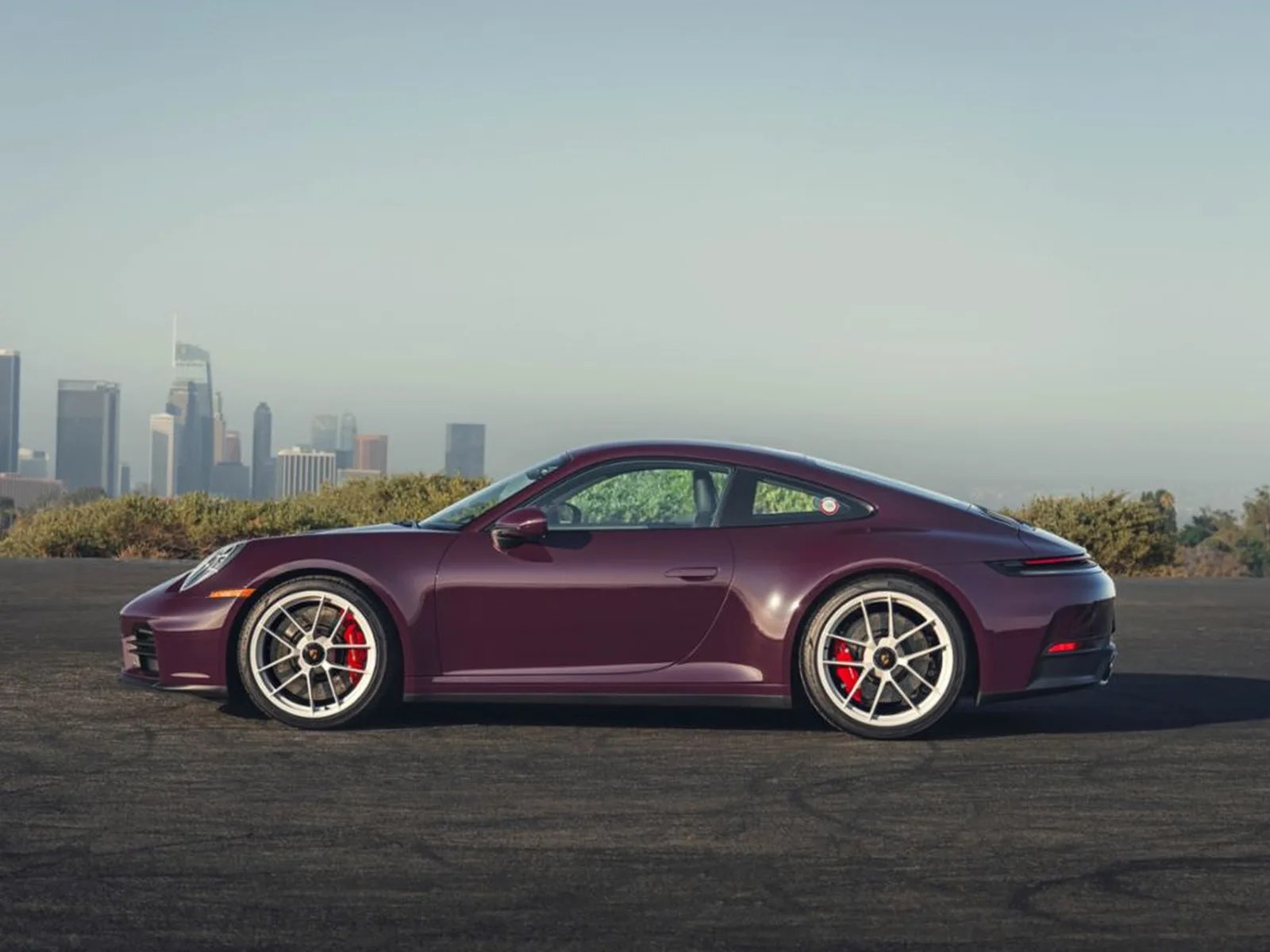 Dark purple Porsche 911 sports car with silver wheels and red brake calipers parked on asphalt with city skyline in background.