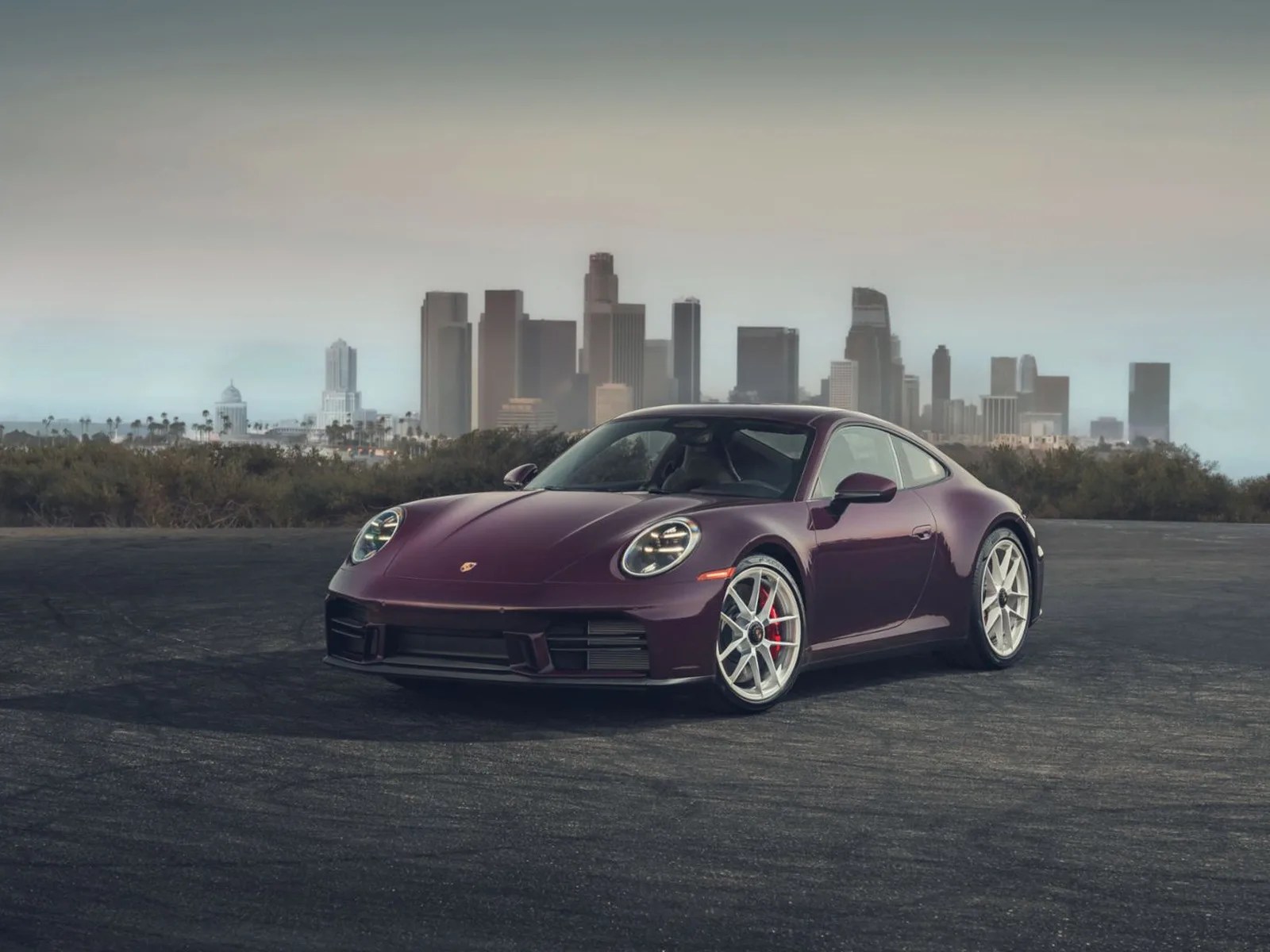Purple Porsche 911 sports car with silver wheels parked on asphalt with a city skyline in the background.