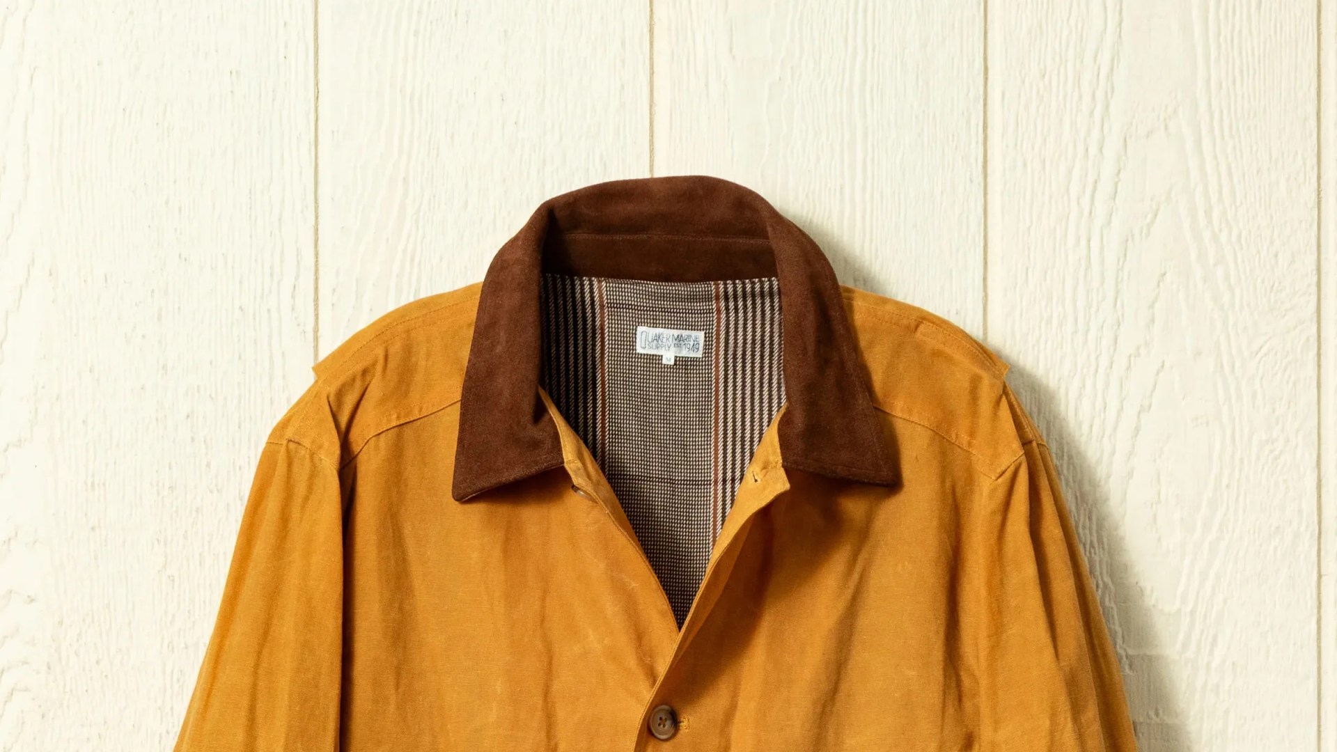 Mustard yellow jacket with dark brown suede collar and plaid inner lining hanging against a white wooden wall.
