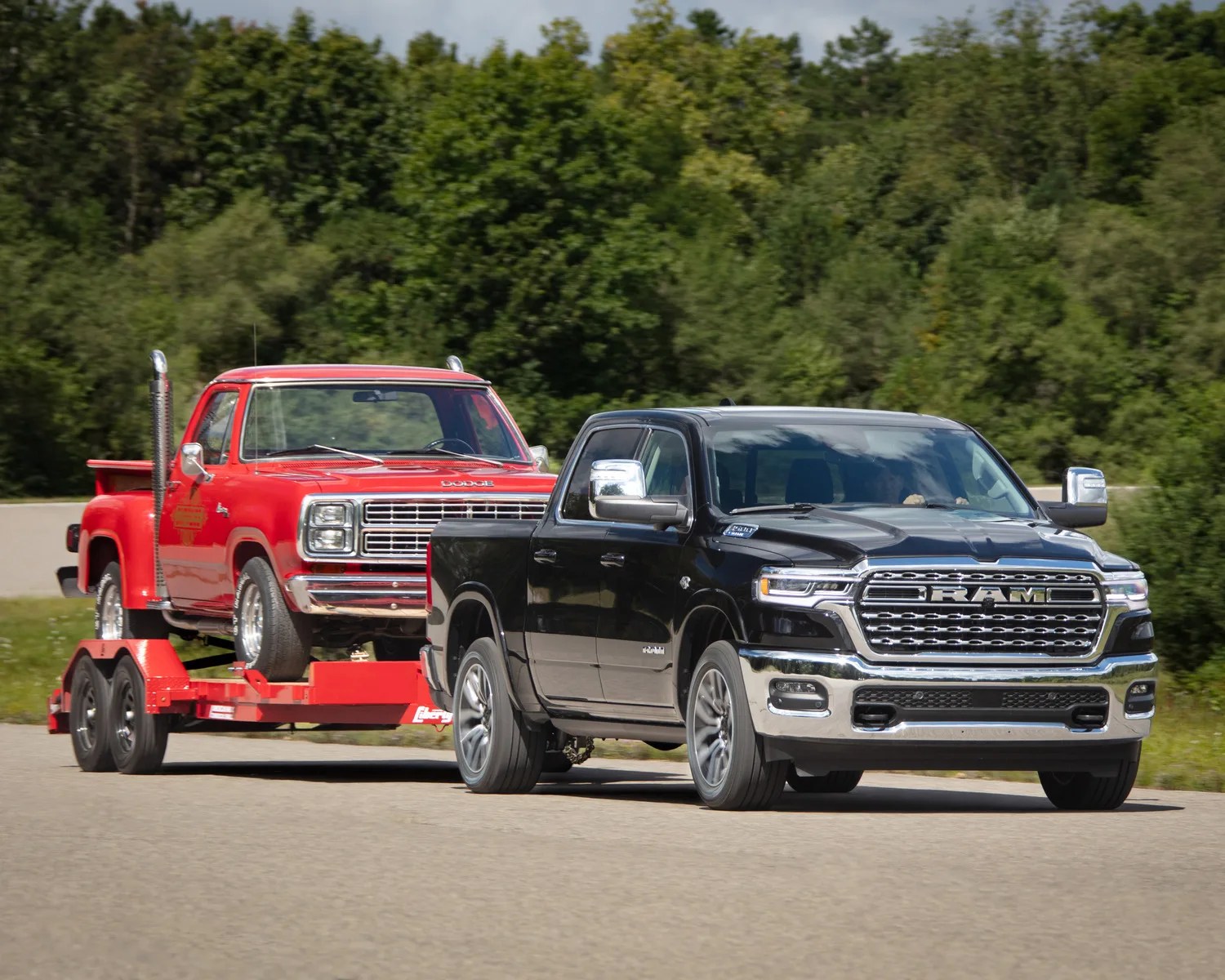 Black RAM pickup truck towing a red vintage Dodge pickup on a red trailer on a road with trees in the background.