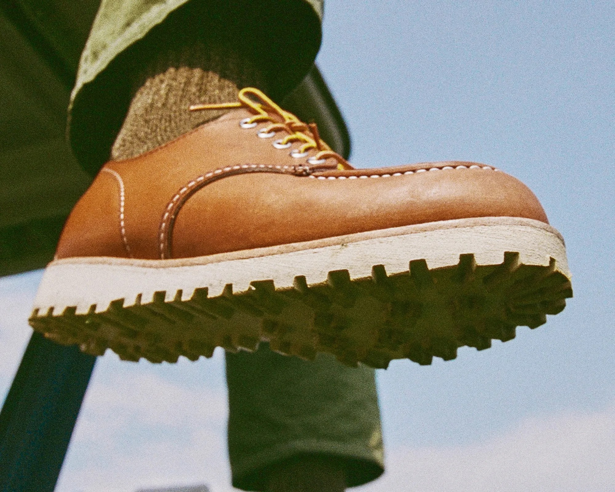 Brown leather shoe with yellow laces and thick white sole worn with green pants and brown socks.