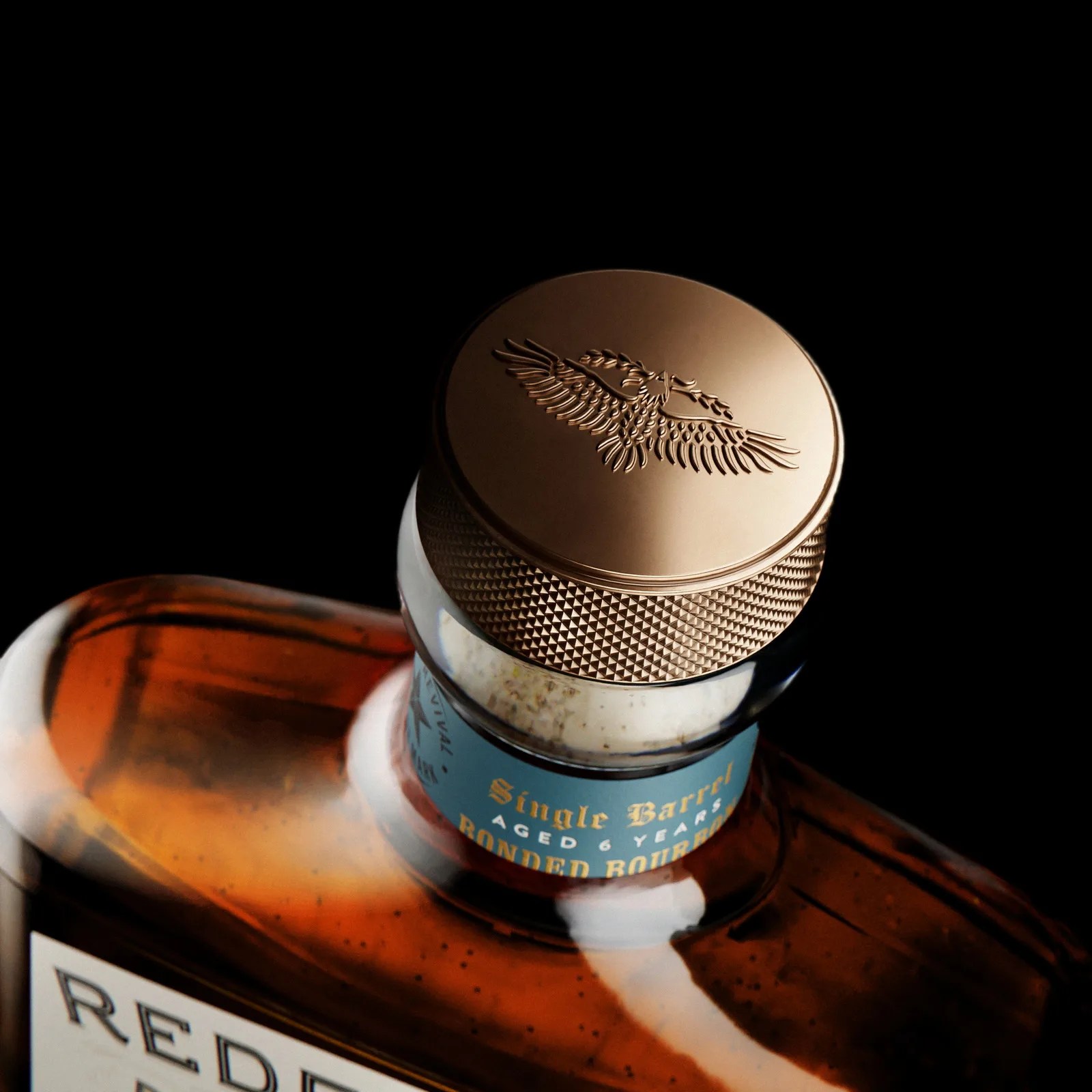 Close-up of a bourbon bottle neck with a textured bronze cap embossed with an eagle holding an olive branch.