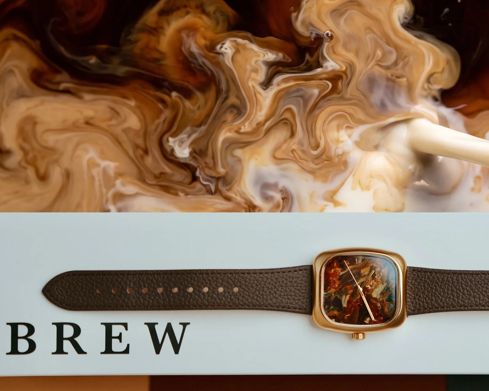 Brown leather strap watch with a gold square case and abstract multicolor dial on a light background with coffee cream swirls.