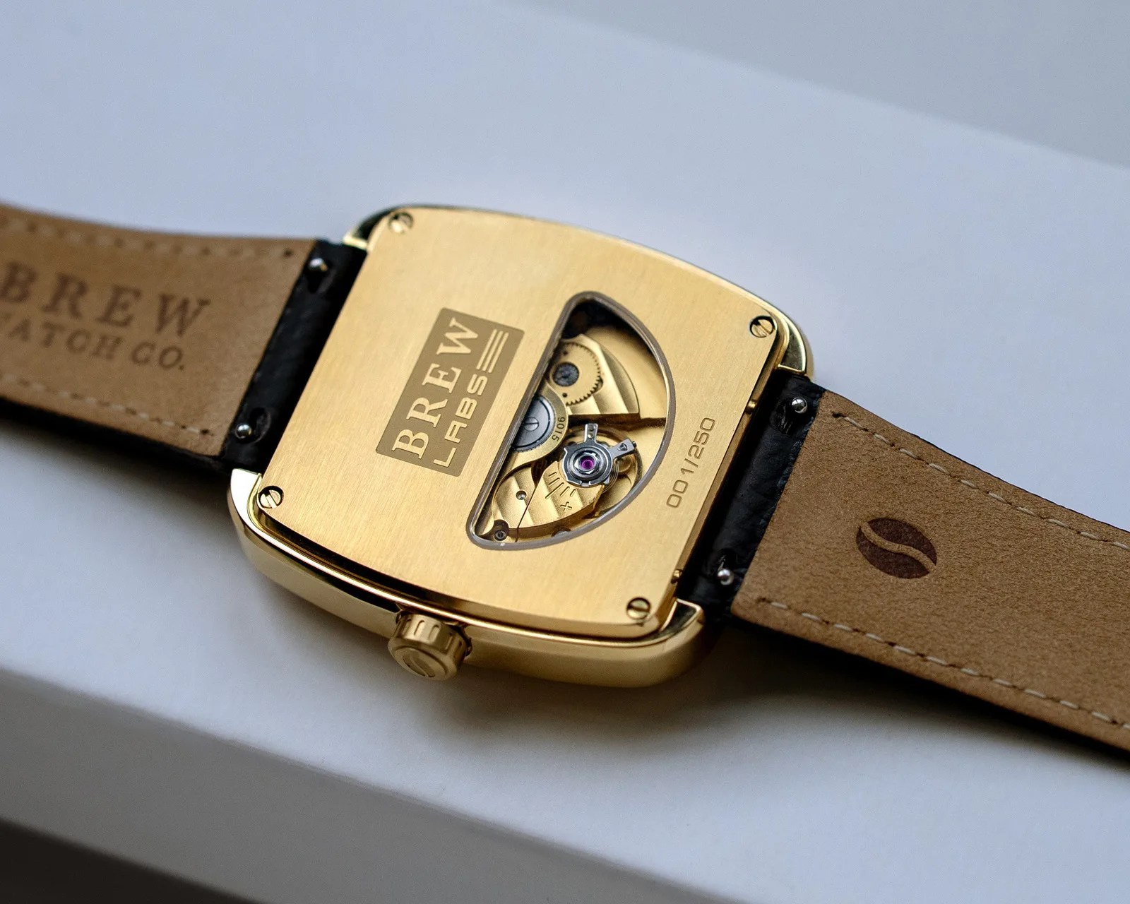 Gold-tone Brew Labs watch back with visible mechanical movement and tan leather strap on a white surface.