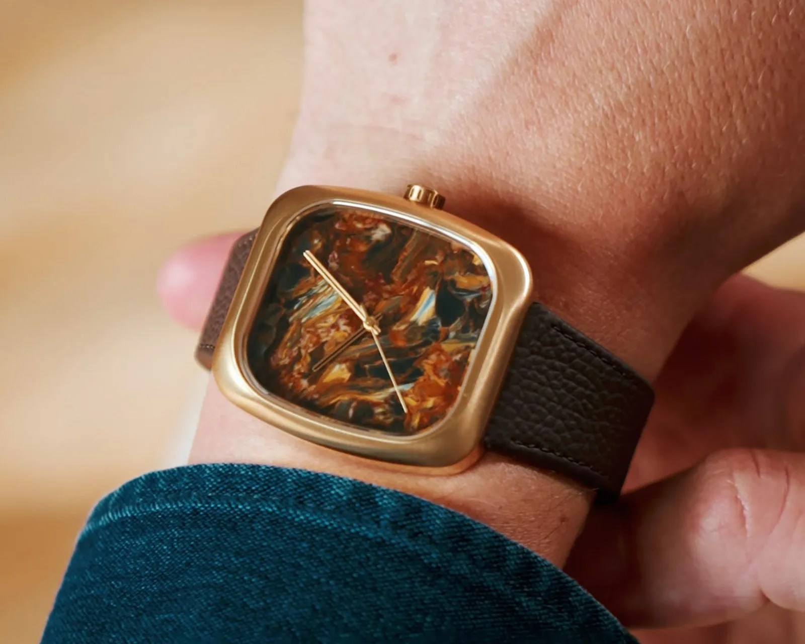 Square gold watch with a marbled brown and black face and a textured dark brown leather strap on a wrist.