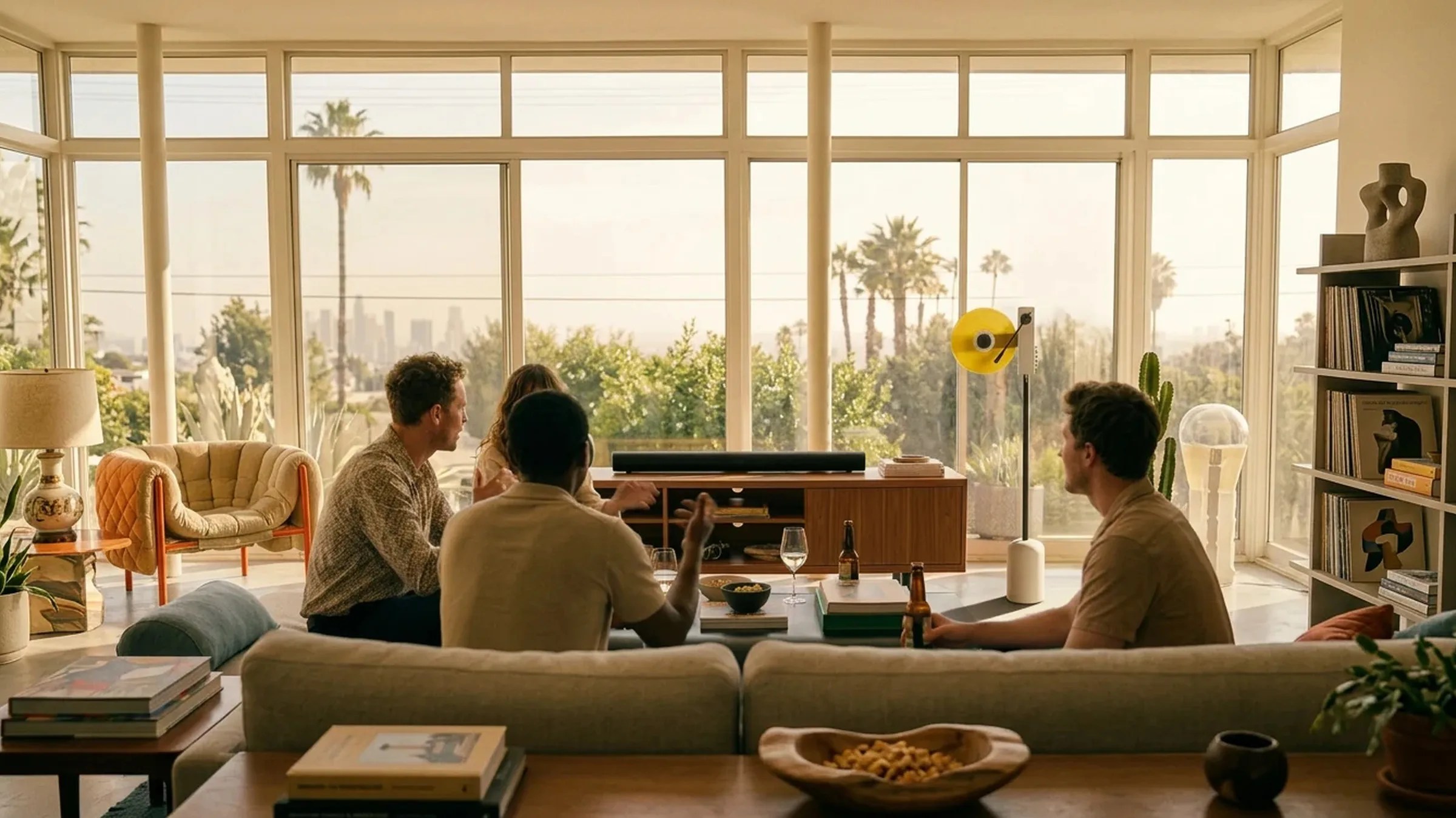 Living room with large windows, beige sofa, wooden coffee table with books and bowl, and four people sitting and talking.