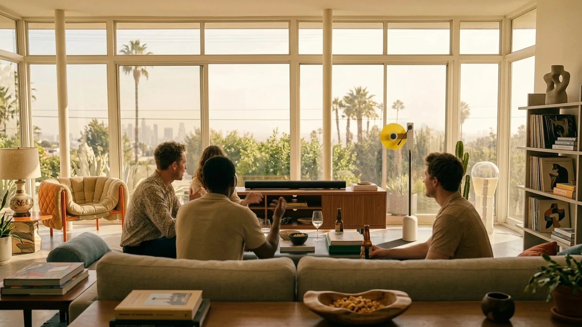 Living room with large windows, beige sofa, wooden coffee table with books and bowl, and four people sitting and talking.