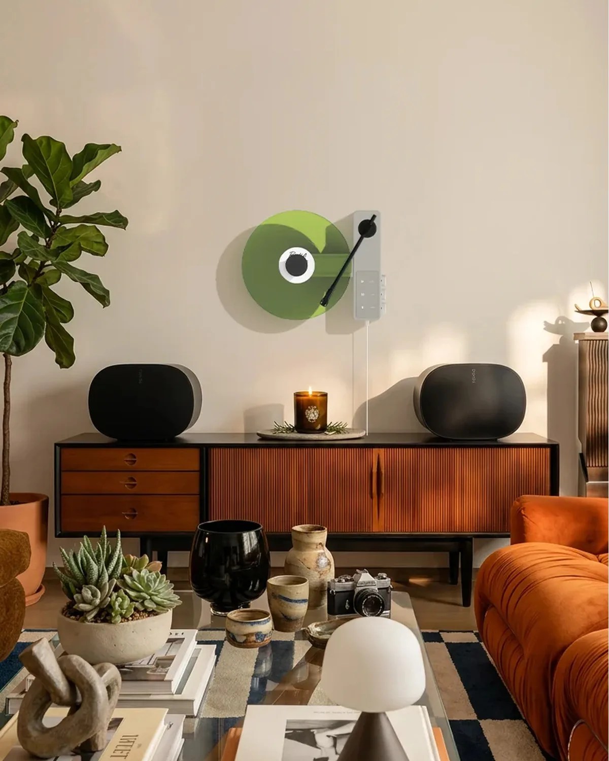 Mid-century modern living room with wooden sideboard, black speakers, green wall-mounted turntable, orange sofa, and glass coffee table with decor.