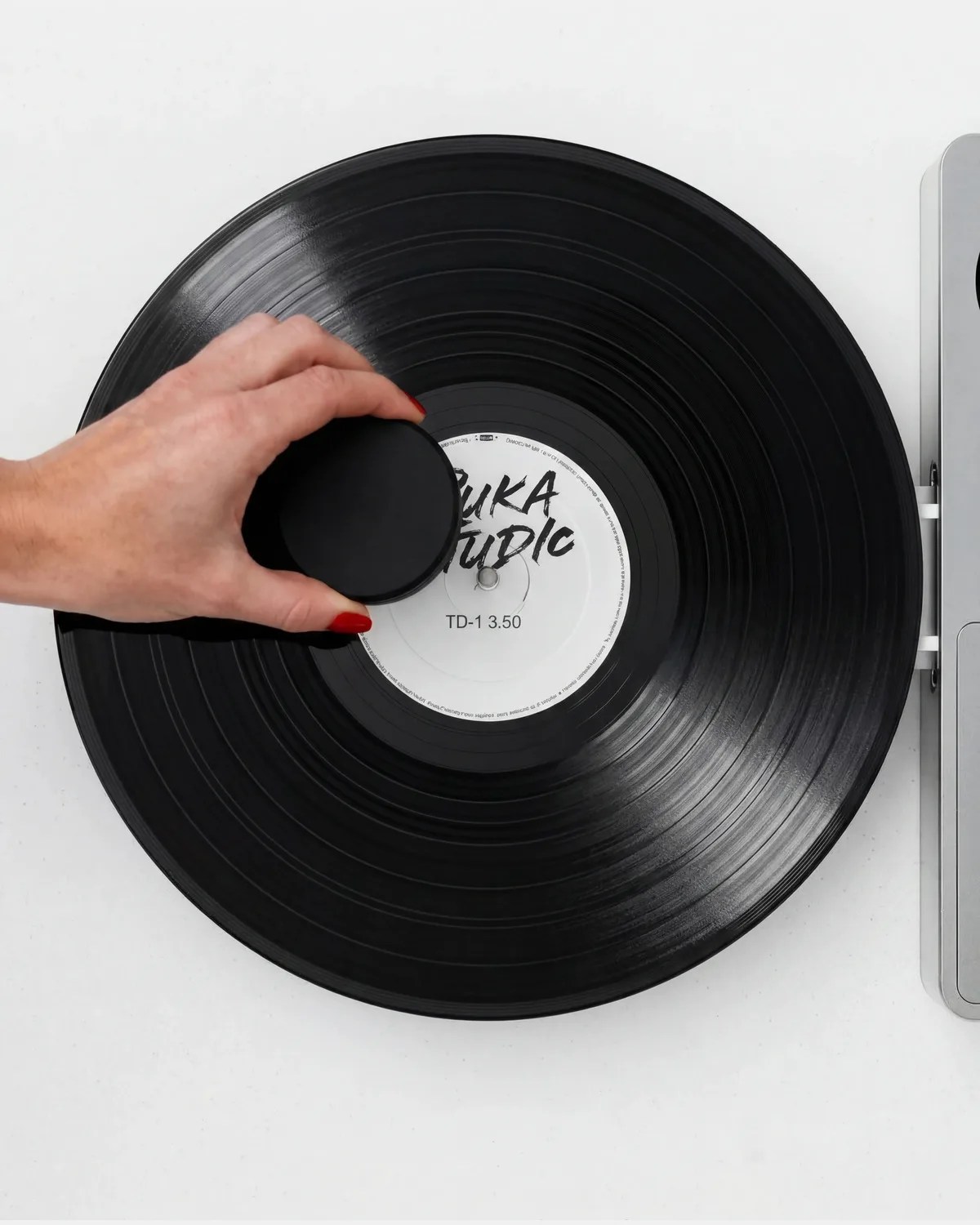 Hand with red nail polish holding a black circular object on a black vinyl record on a white surface.